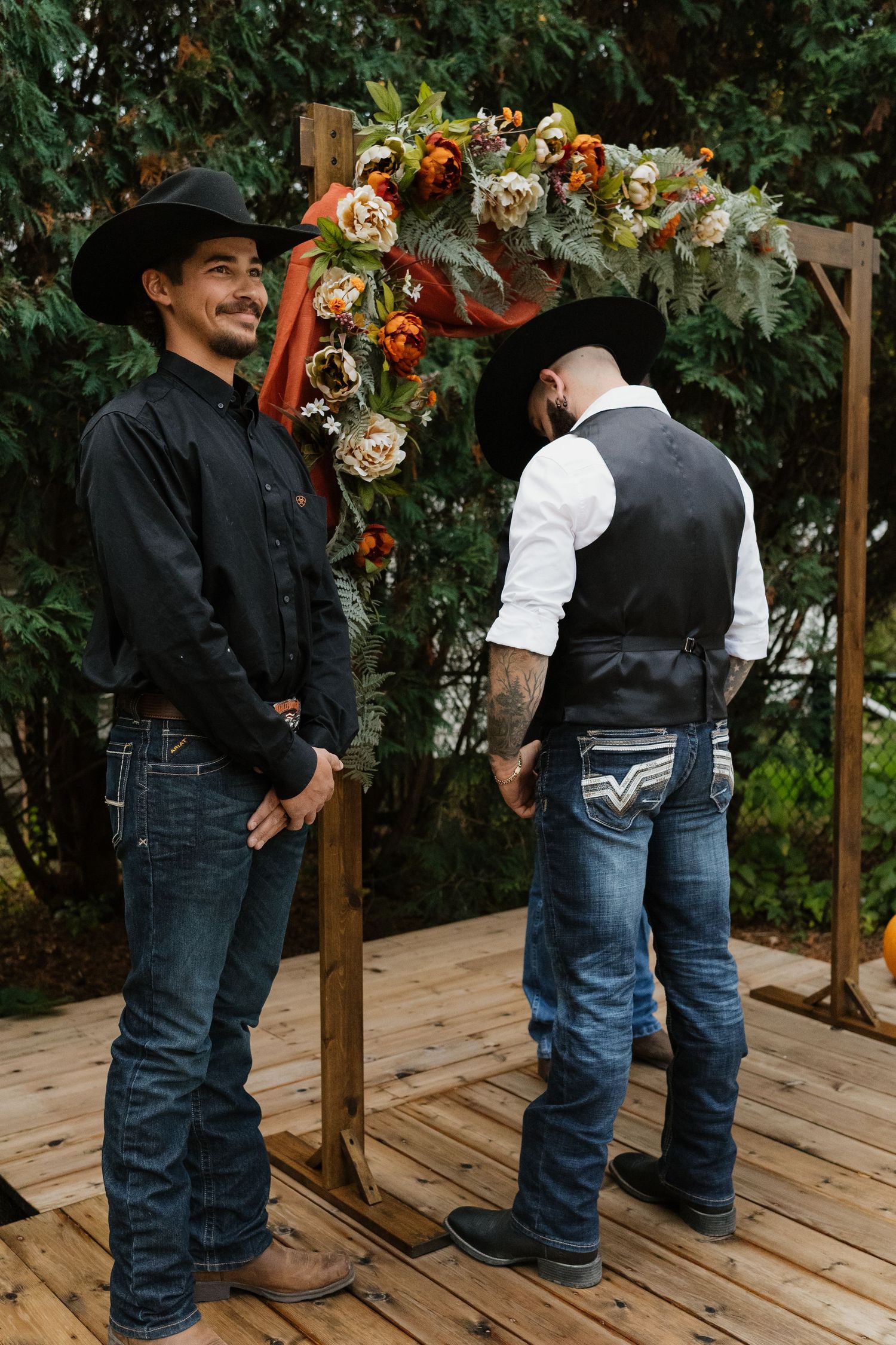 Two groomsmen in black cowboy hats and jeans stand at a rustic floral wedding arch.