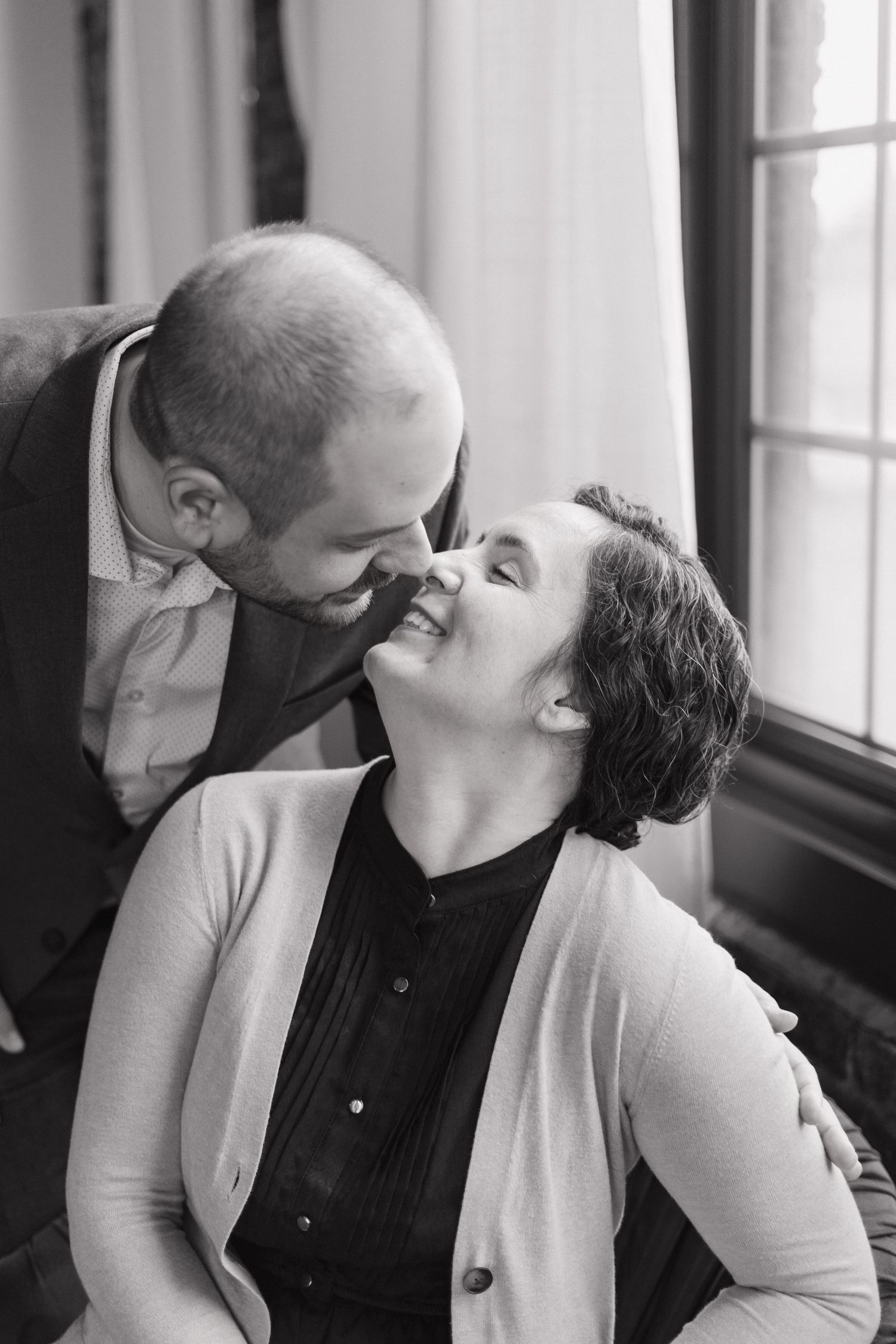 Black and white photo of man leaning down to kiss smiling seated woman by window.