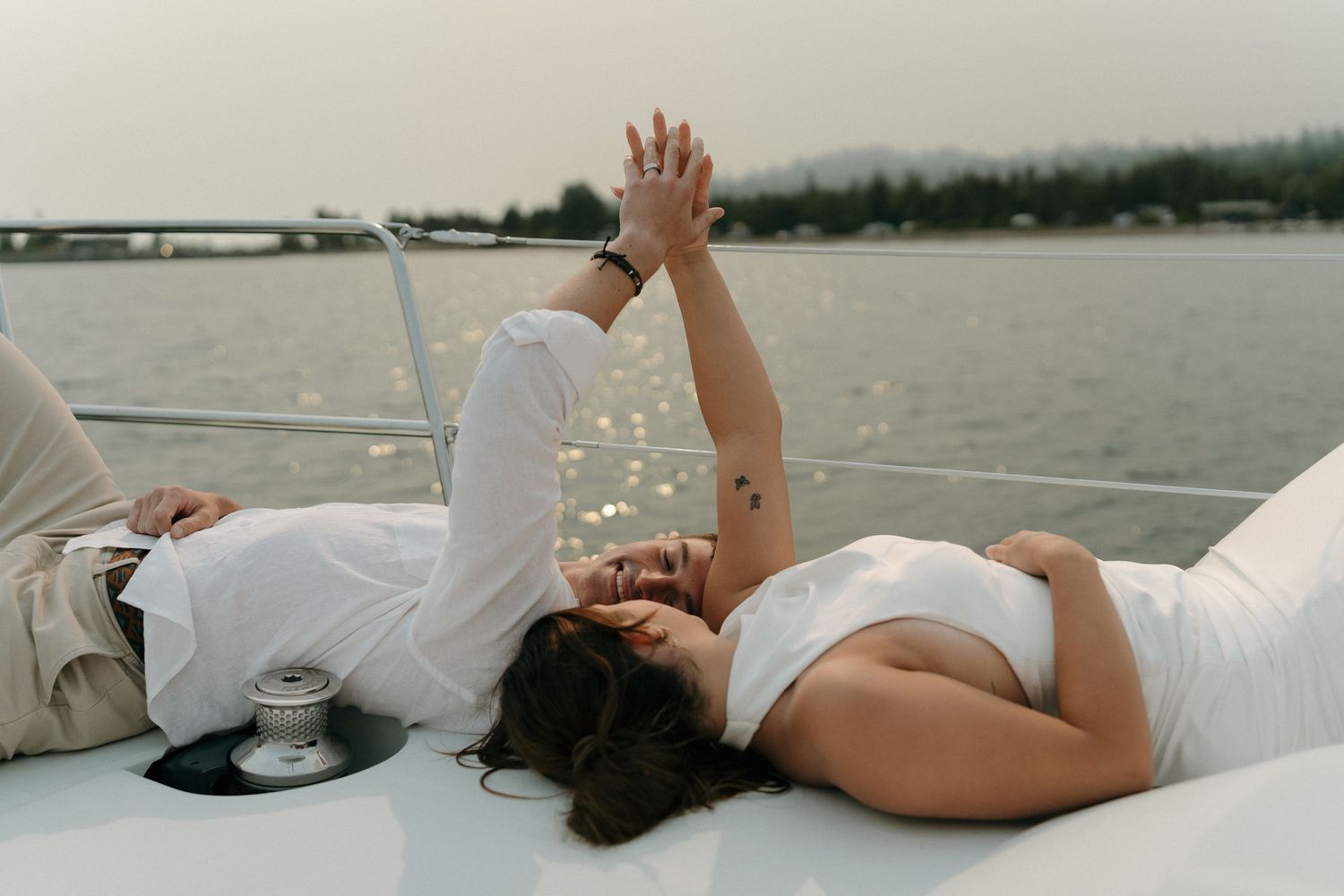 Couple lying on a yacht deck holding hands, dressed in white, with calm ocean water in the background.