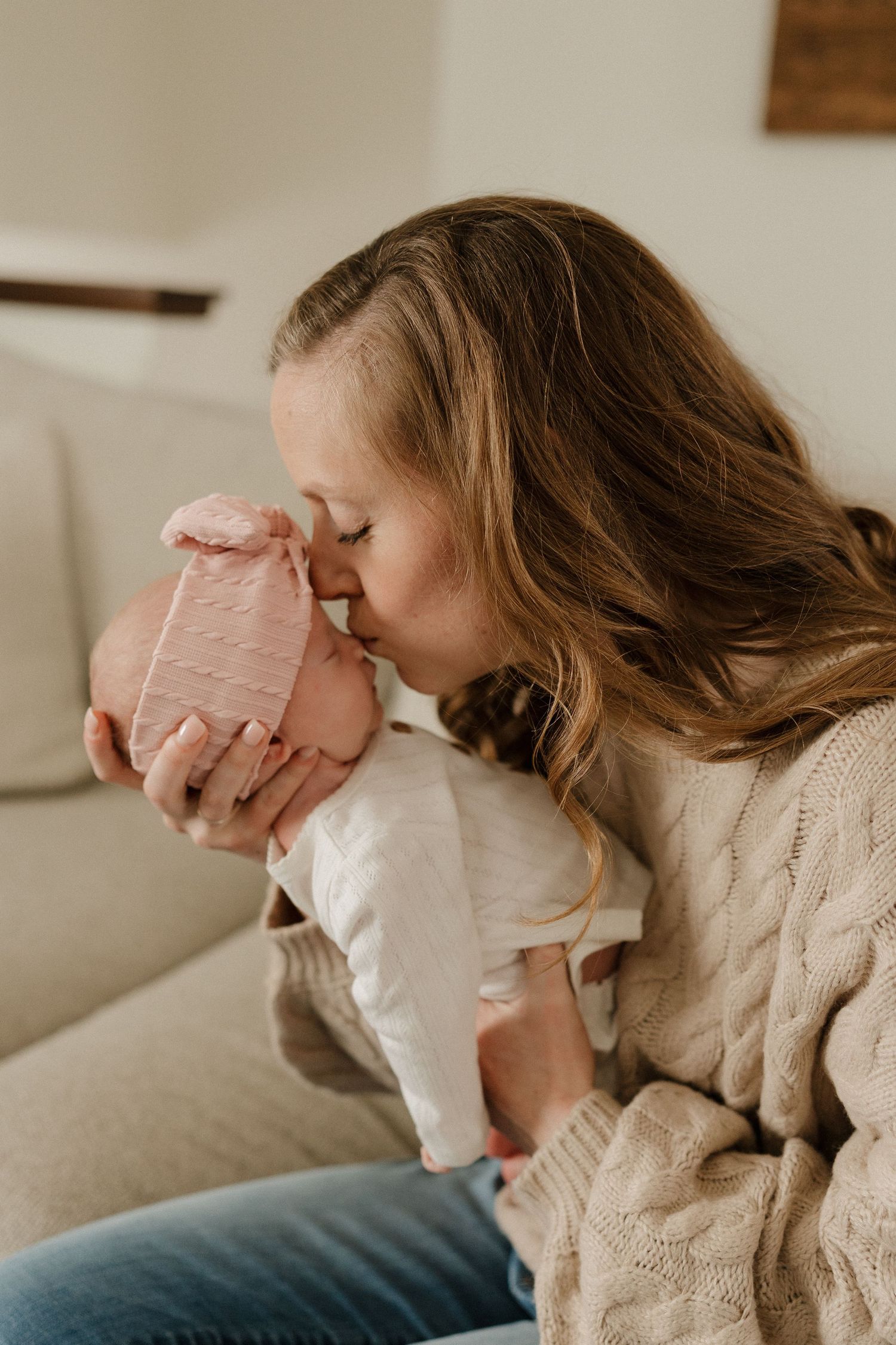 A mother tenderly kisses her newborn baby wearing a pink hat while sitting on a couch.