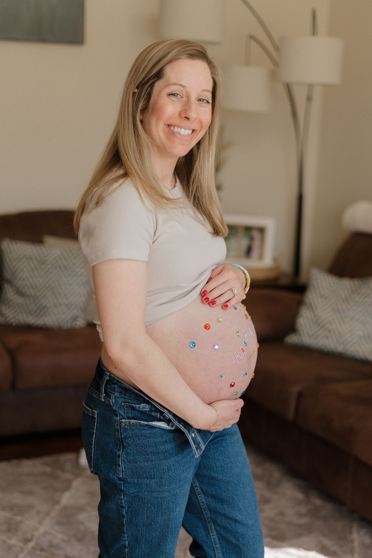 Smiling pregnant woman in jeans and gray top holding baby bump decorated with colorful gems indoors.