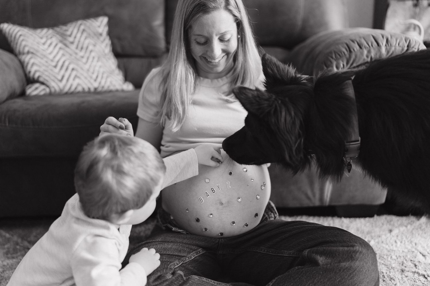 Black and white photo of pregnant woman smiling at toddler with dog beside her on living room floor.