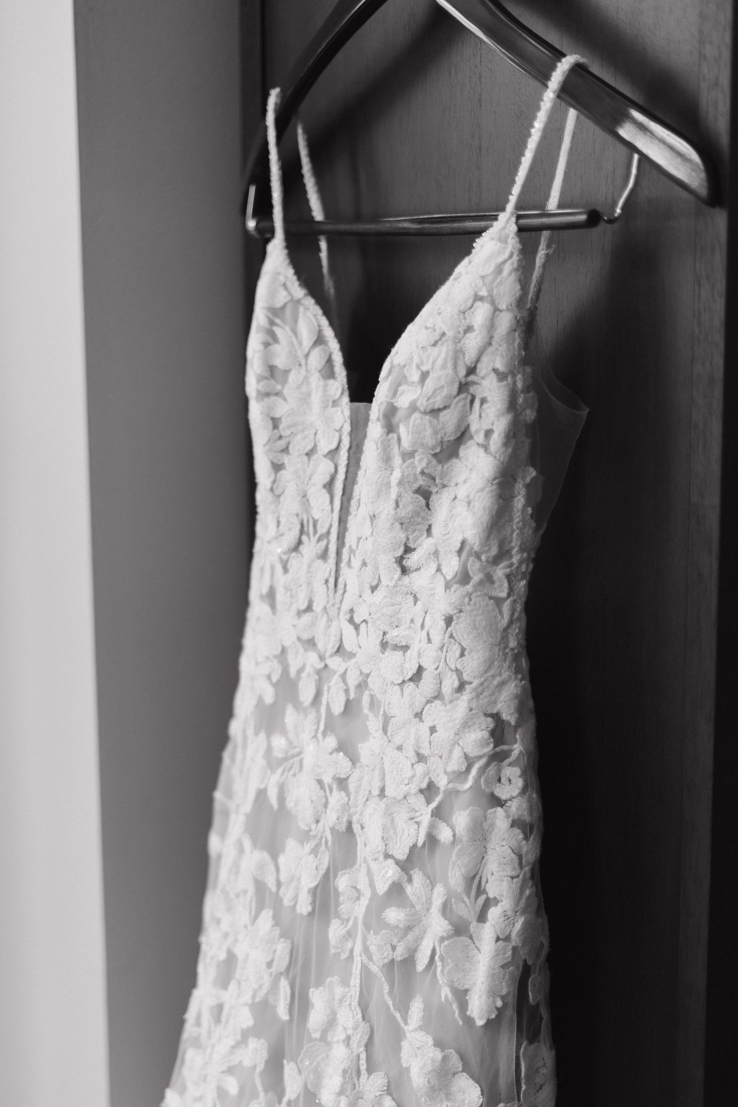 Black and white photo of an elegant lace wedding dress with floral appliques hanging on a wooden hanger.