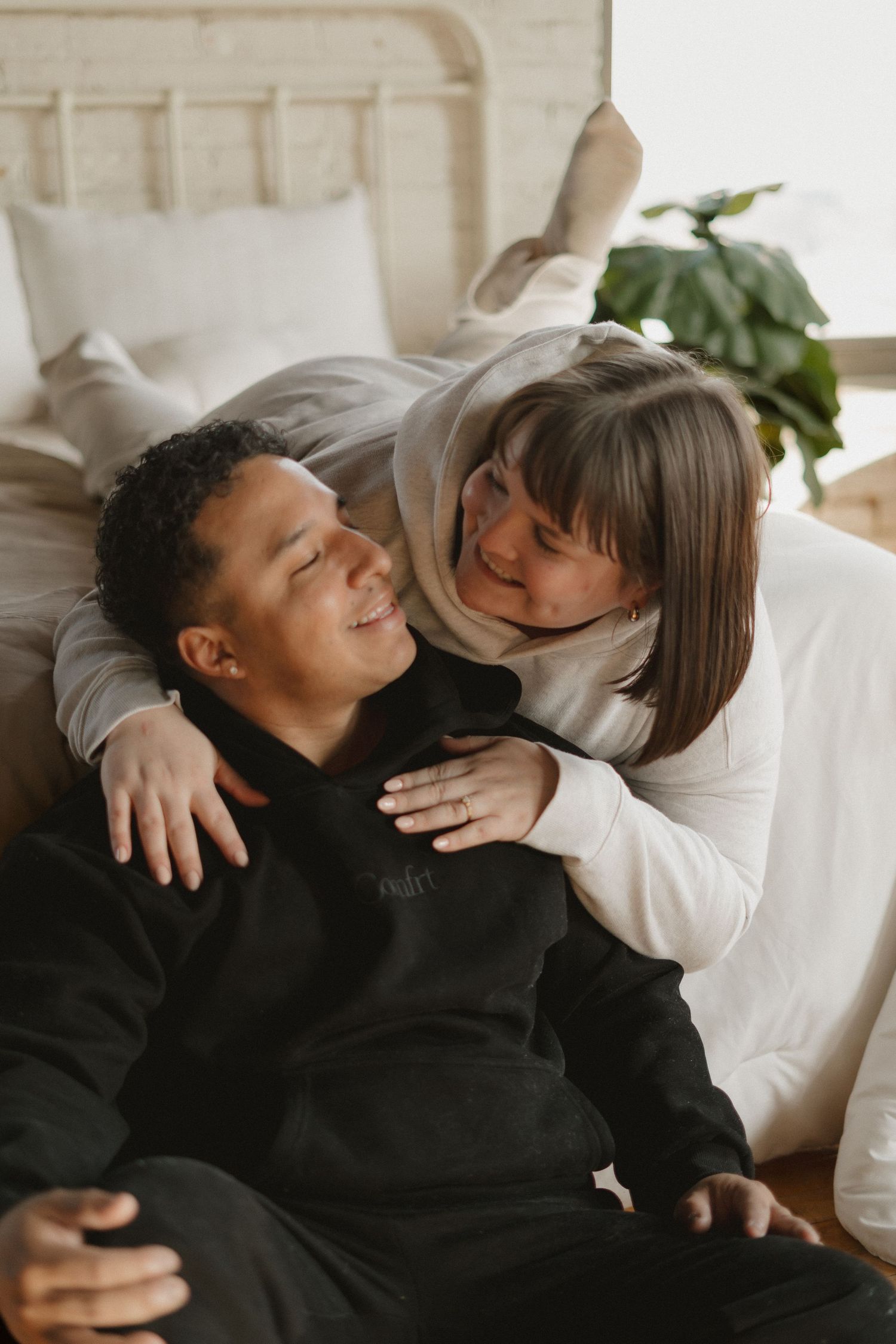 Happy interracial couple embracing and smiling together on bedroom floor, wearing casual loungewear.