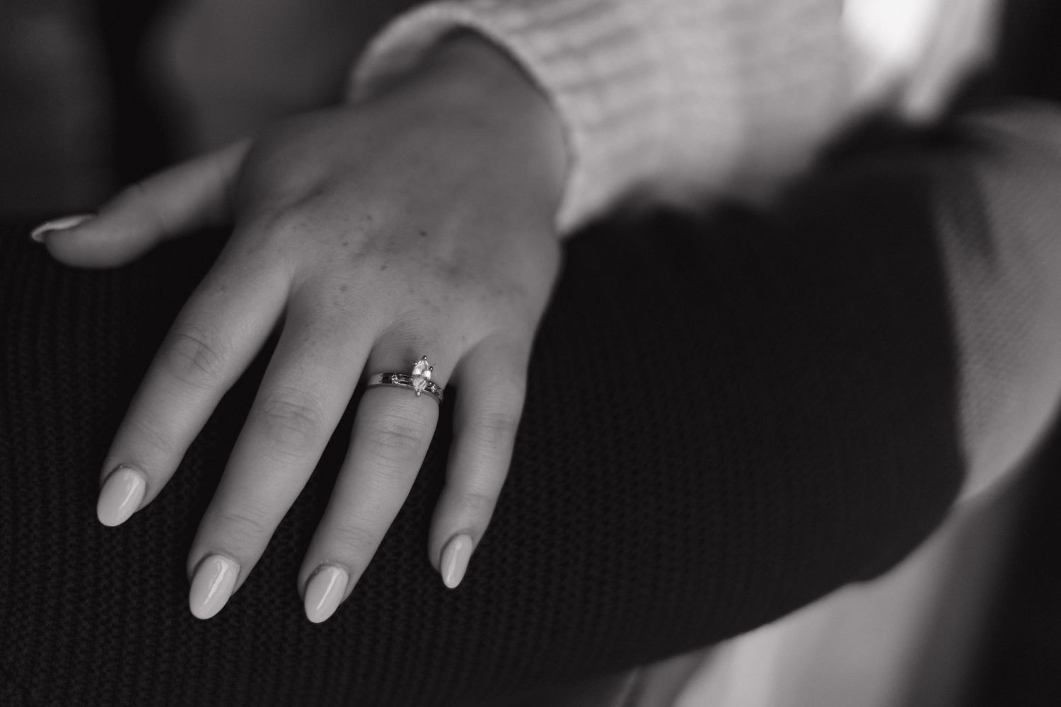 Black and white photo of a woman's hand wearing a diamond engagement ring, resting on dark fabric.