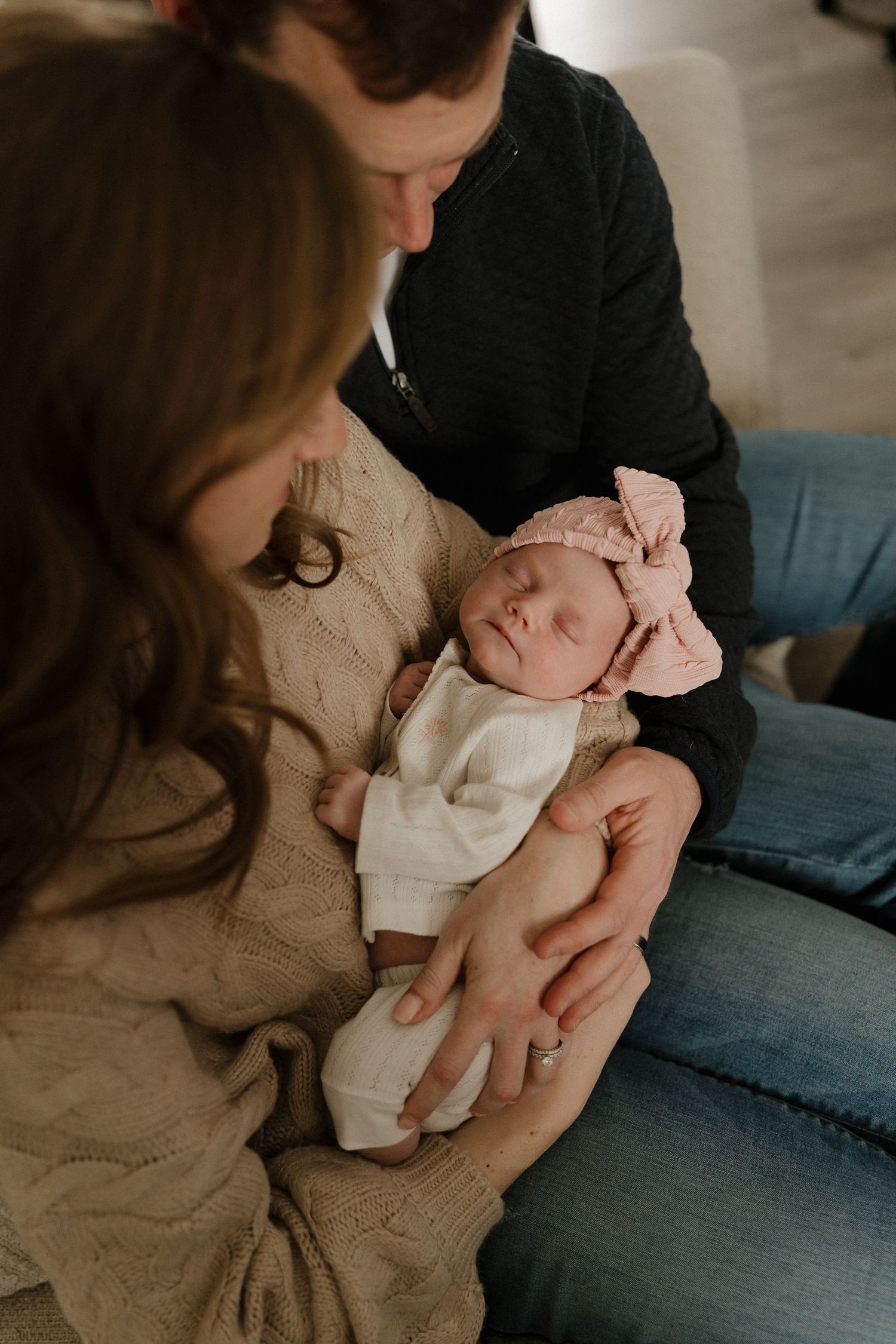 Parents lovingly gaze at their sleeping newborn baby girl wearing a pink bow headband and white outfit.