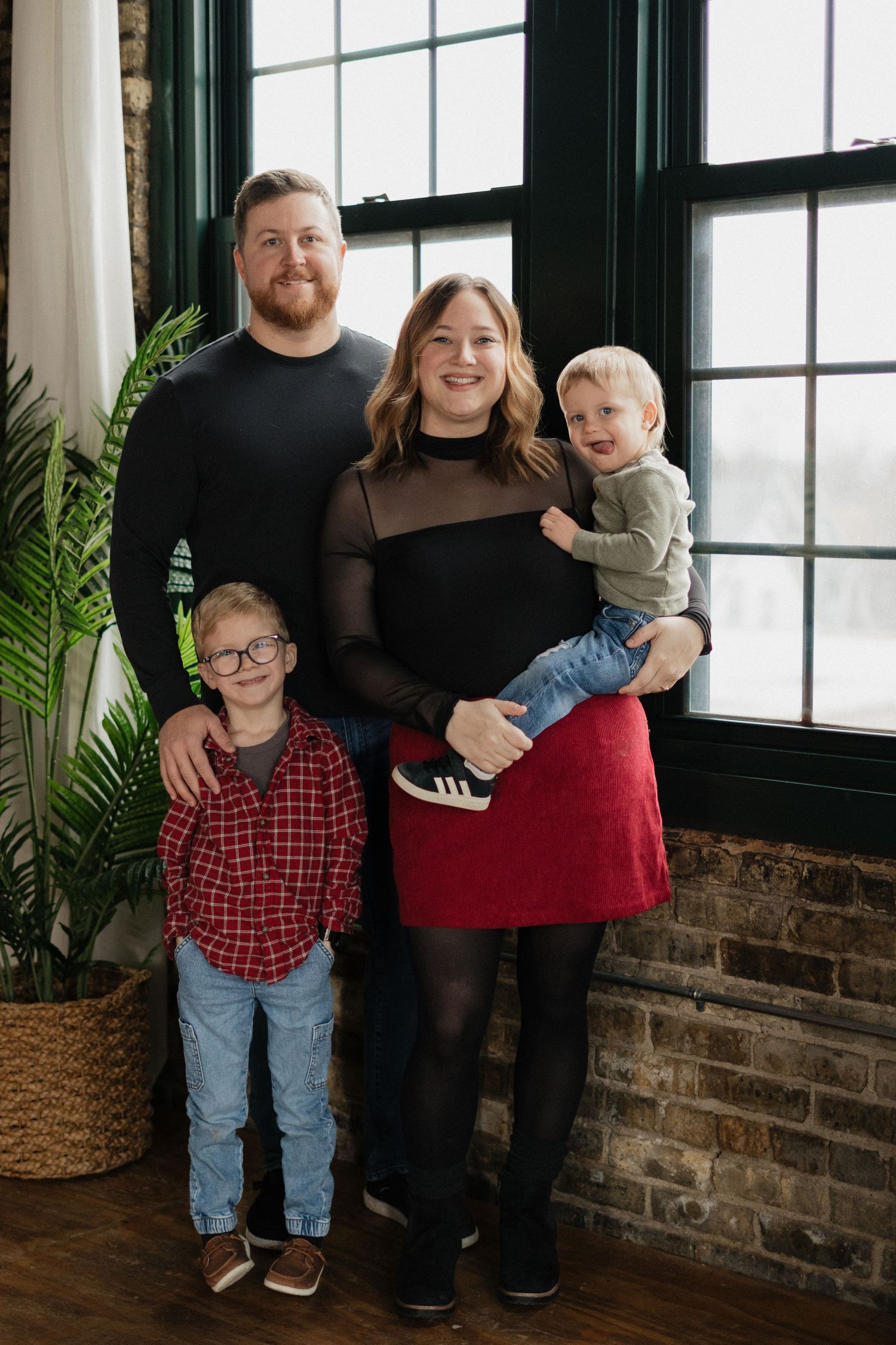 Family of four poses indoors by large windows, parents with two young boys in coordinated outfits.