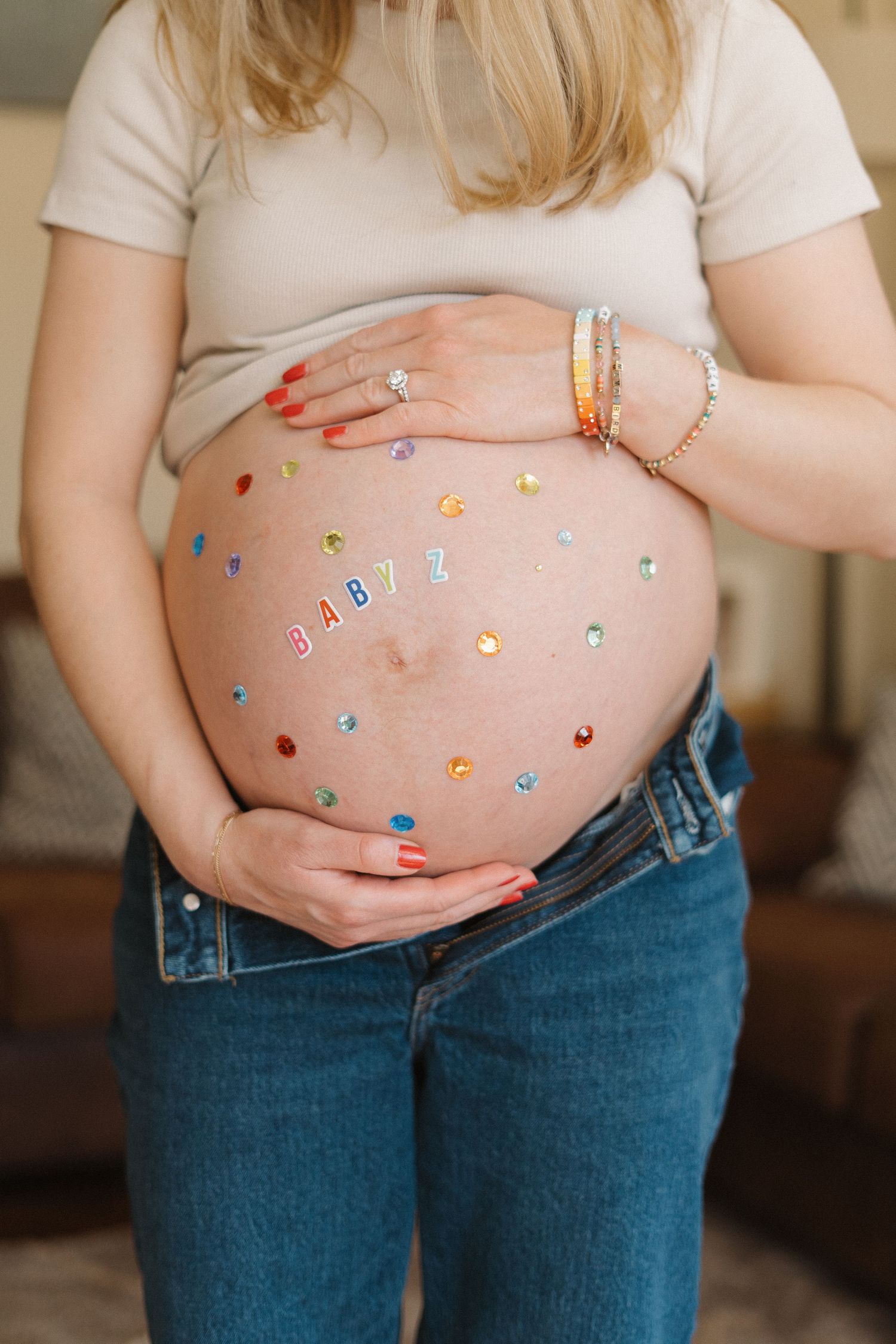 Pregnant woman in jeans displays decorated baby bump with colorful gems and BABY letters spelling message.