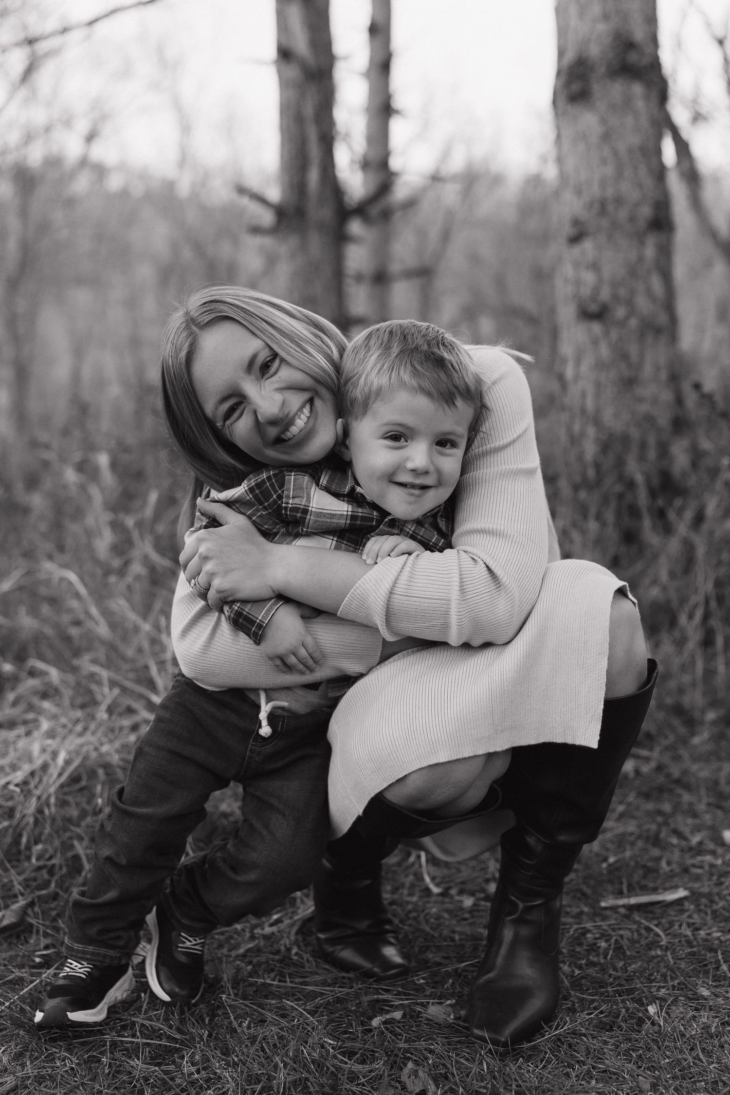 Black and white photo of blonde mother hugging young son in plaid shirt in woodland setting.