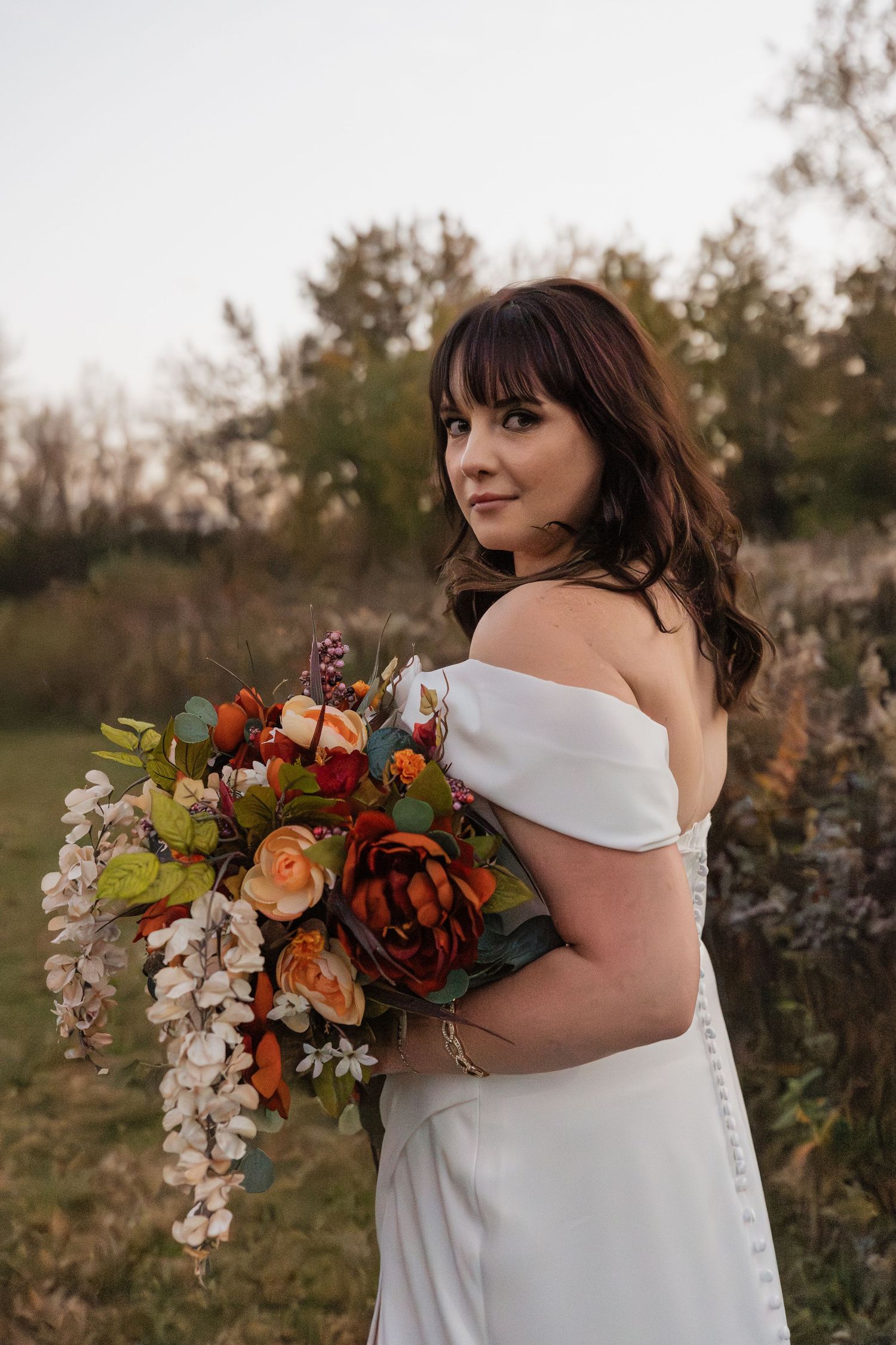 Bride in off-shoulder white gown looks back holding lush autumn bouquet in outdoor fall setting.