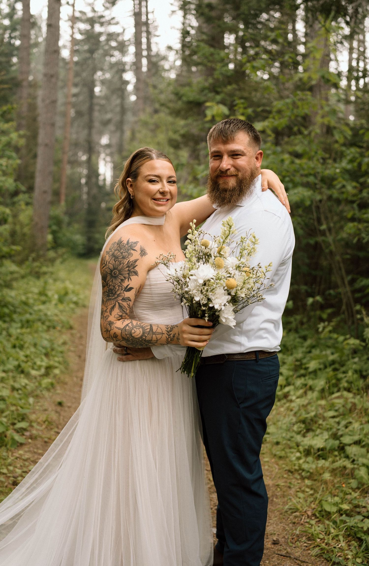 A tattooed bride in a white tulle gown holds a wildflower bouquet beside her bearded groom in a forest setting.