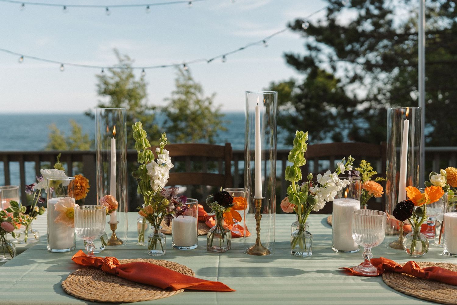 Elegant outdoor wedding table with colorful floral arrangements, candles, and ocean view backdrop under string lights.