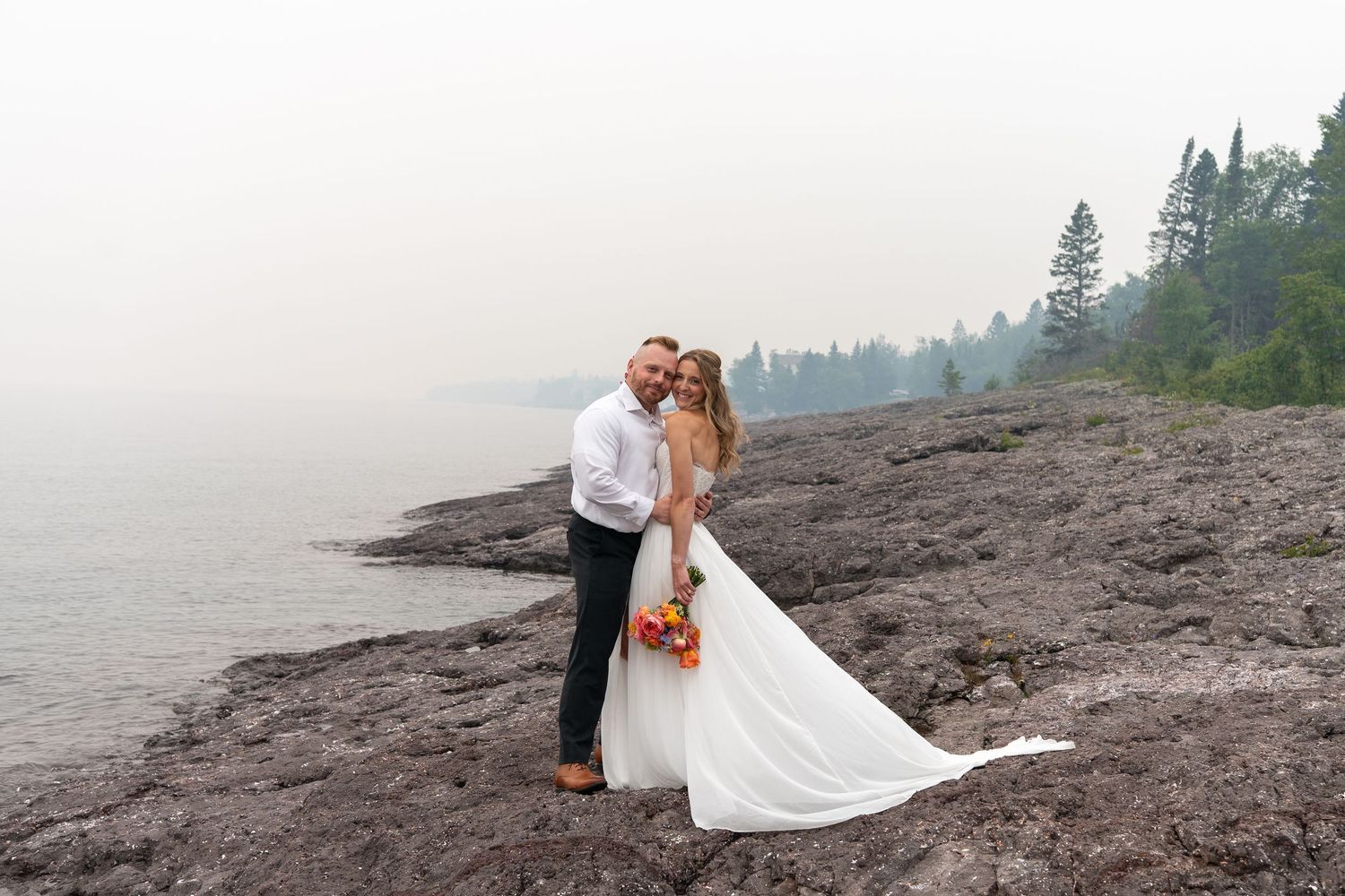 Bride and groom embrace on rocky Lake Superior shoreline in misty forest setting, bride holding vibrant orange bouquet.
