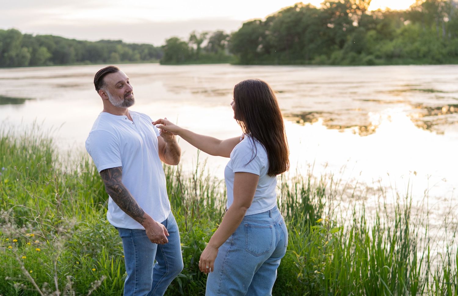 Couple in matching white tees and jeans share a tender moment by a serene river surrounded by lush greenery.