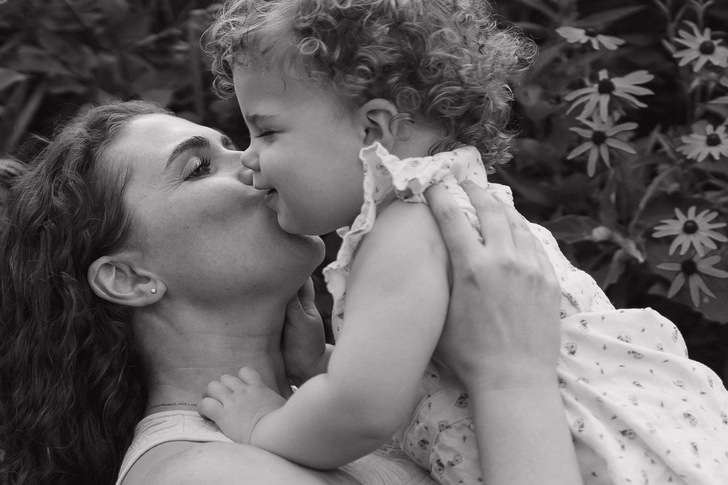 Black and white photo of a mother and curly-haired toddler sharing a sweet kiss outdoors.