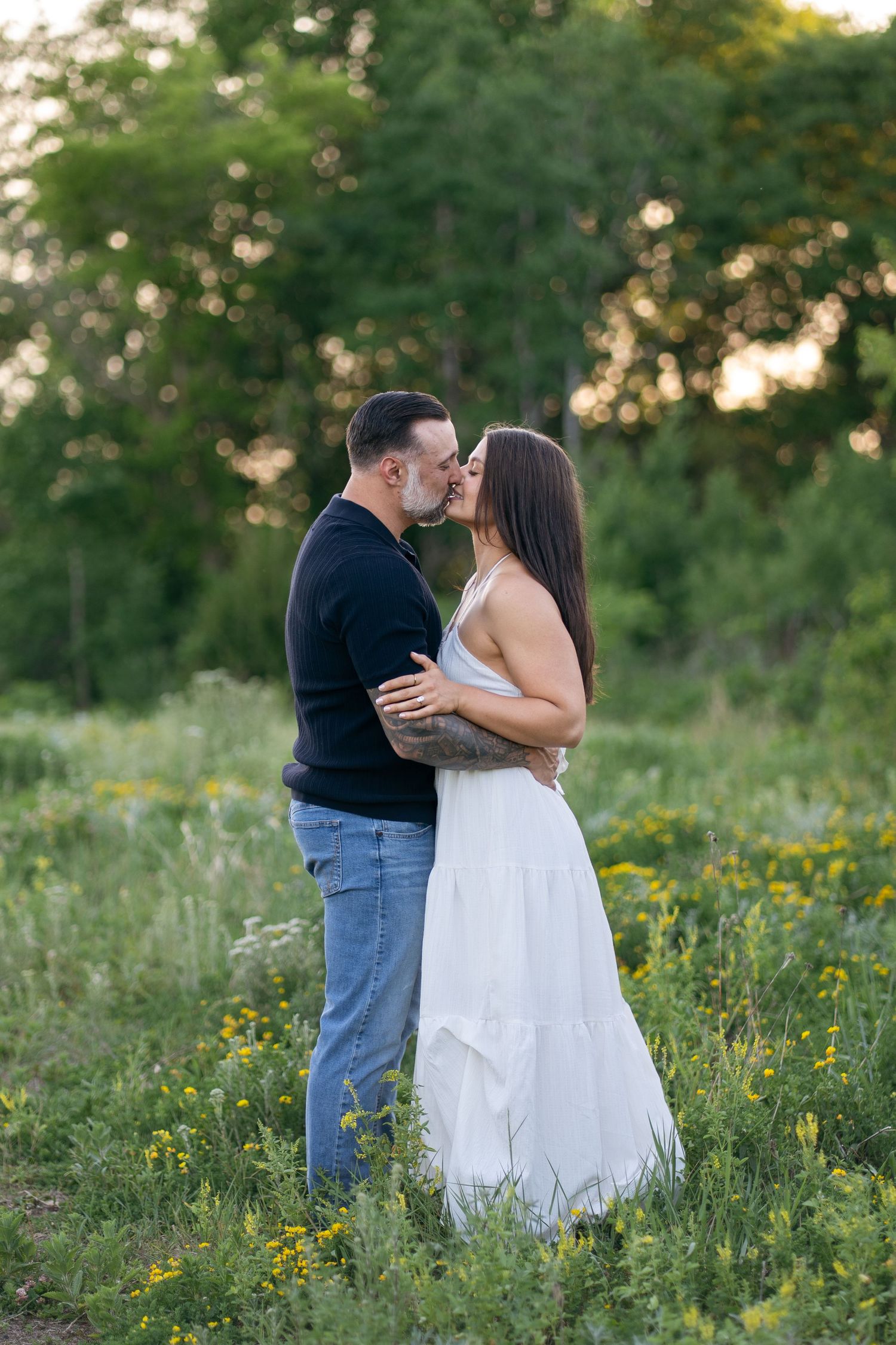 Couple sharing a romantic kiss in a lush green field with yellow wildflowers, man in black shirt and woman in white dress.