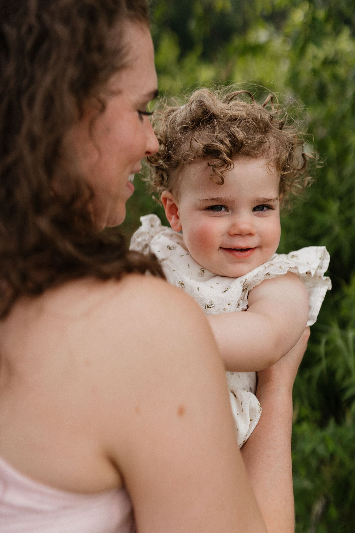 A curly-haired toddler in a white floral dress smiles while being held by her mother outdoors.