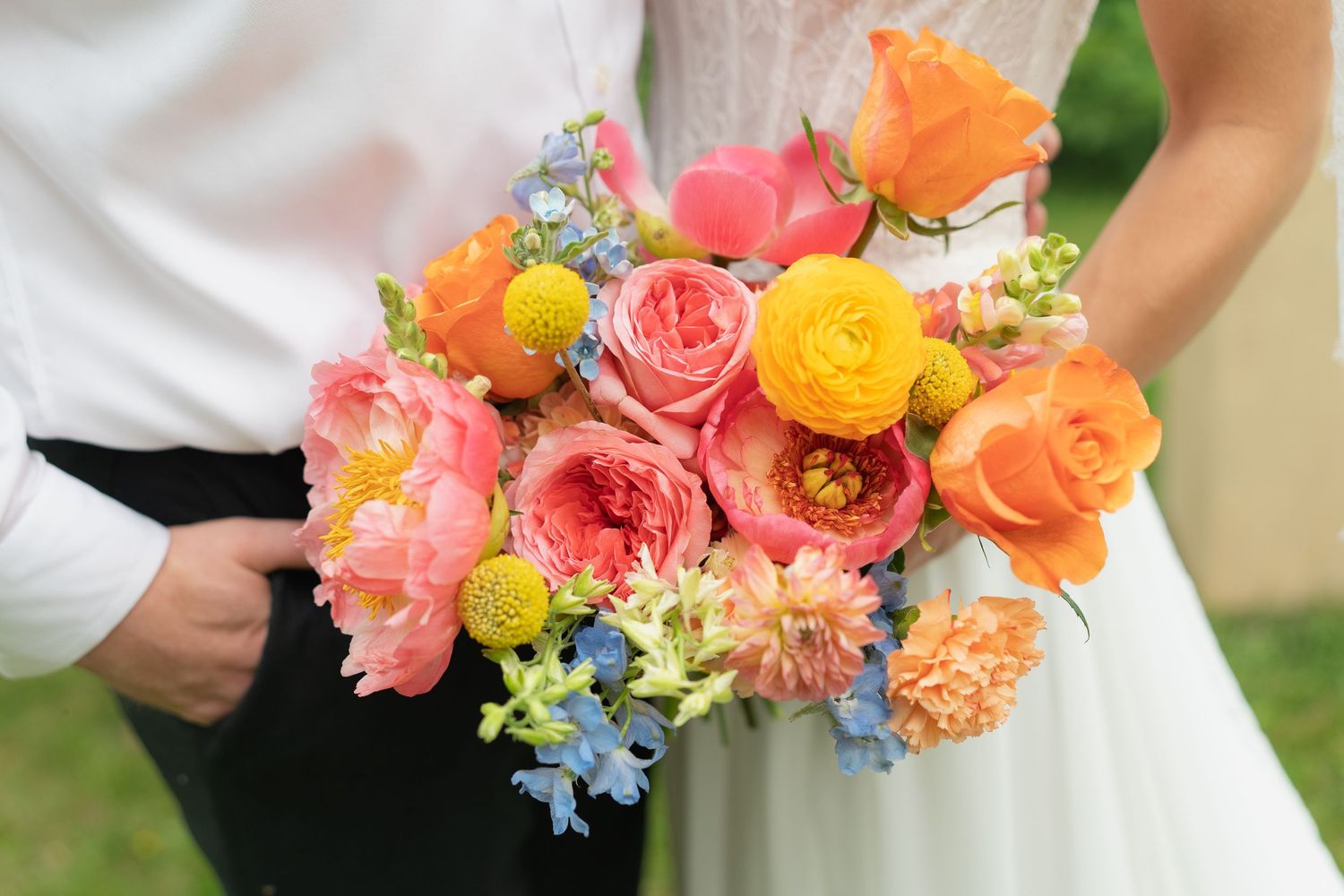 Colorful wedding bouquet featuring pink peonies, orange roses, yellow ranunculus, and blue delphinium held by bride and groom.