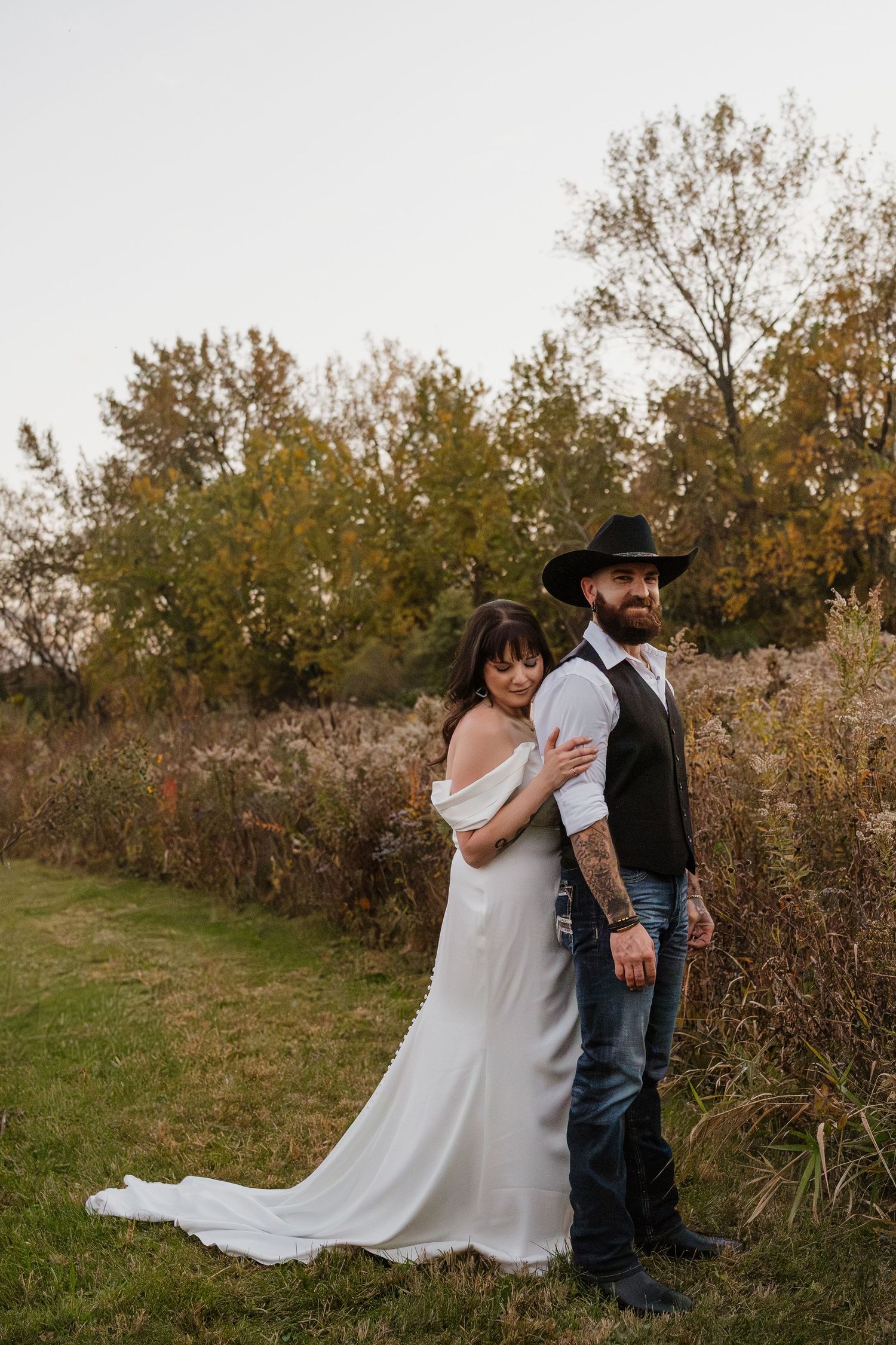 Bride in elegant white gown leans against groom in cowboy hat standing in an autumn landscape.