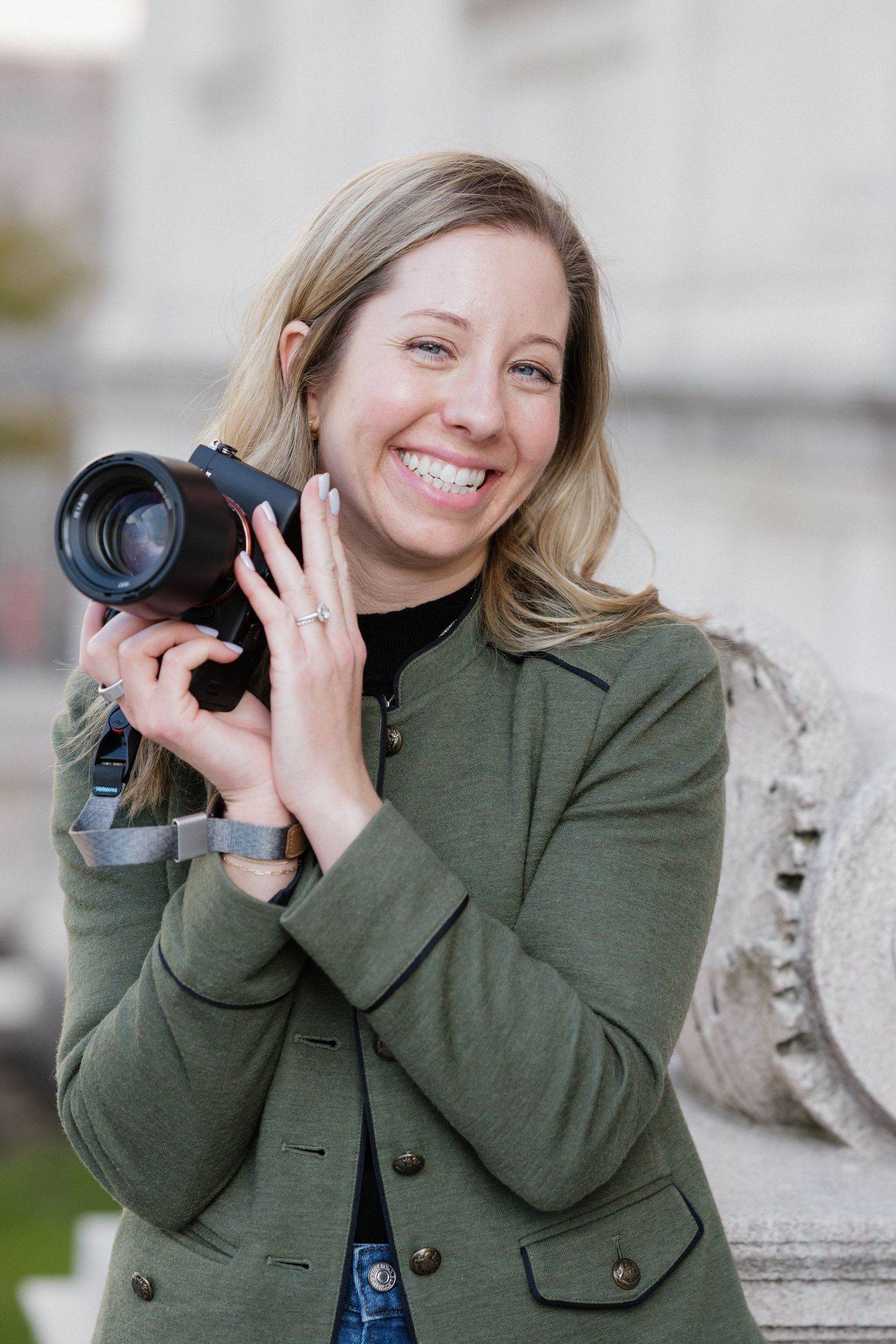 Smiling blonde female photographer in green coat holding camera outdoors near a stone building column.