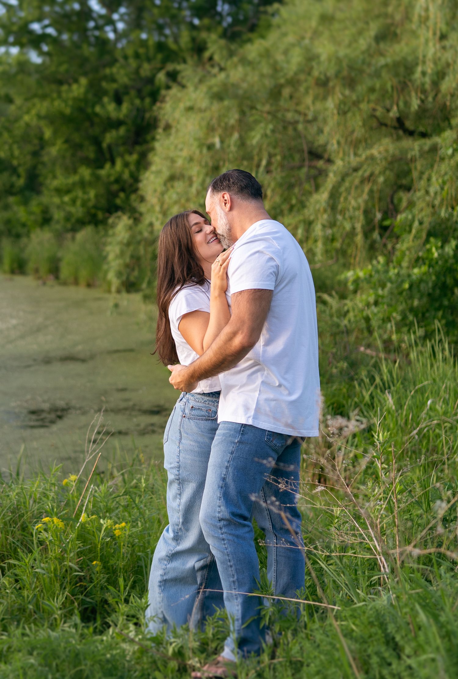A couple in matching white shirts and jeans share a romantic embrace near a lush green pond outdoors.