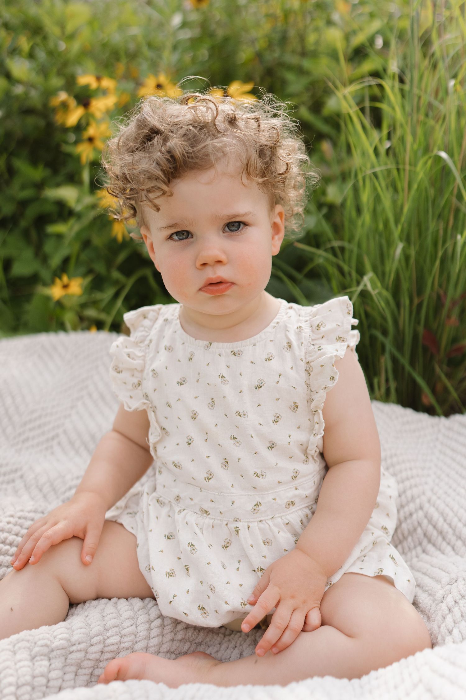 Curly-haired toddler girl in floral romper sits on white blanket near yellow garden flowers.
