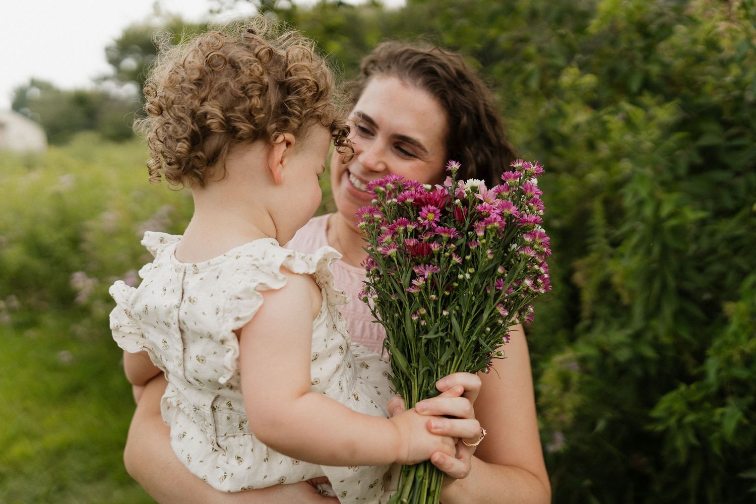 A mother holds her toddler and a bouquet of pink flowers while they share a tender moment.