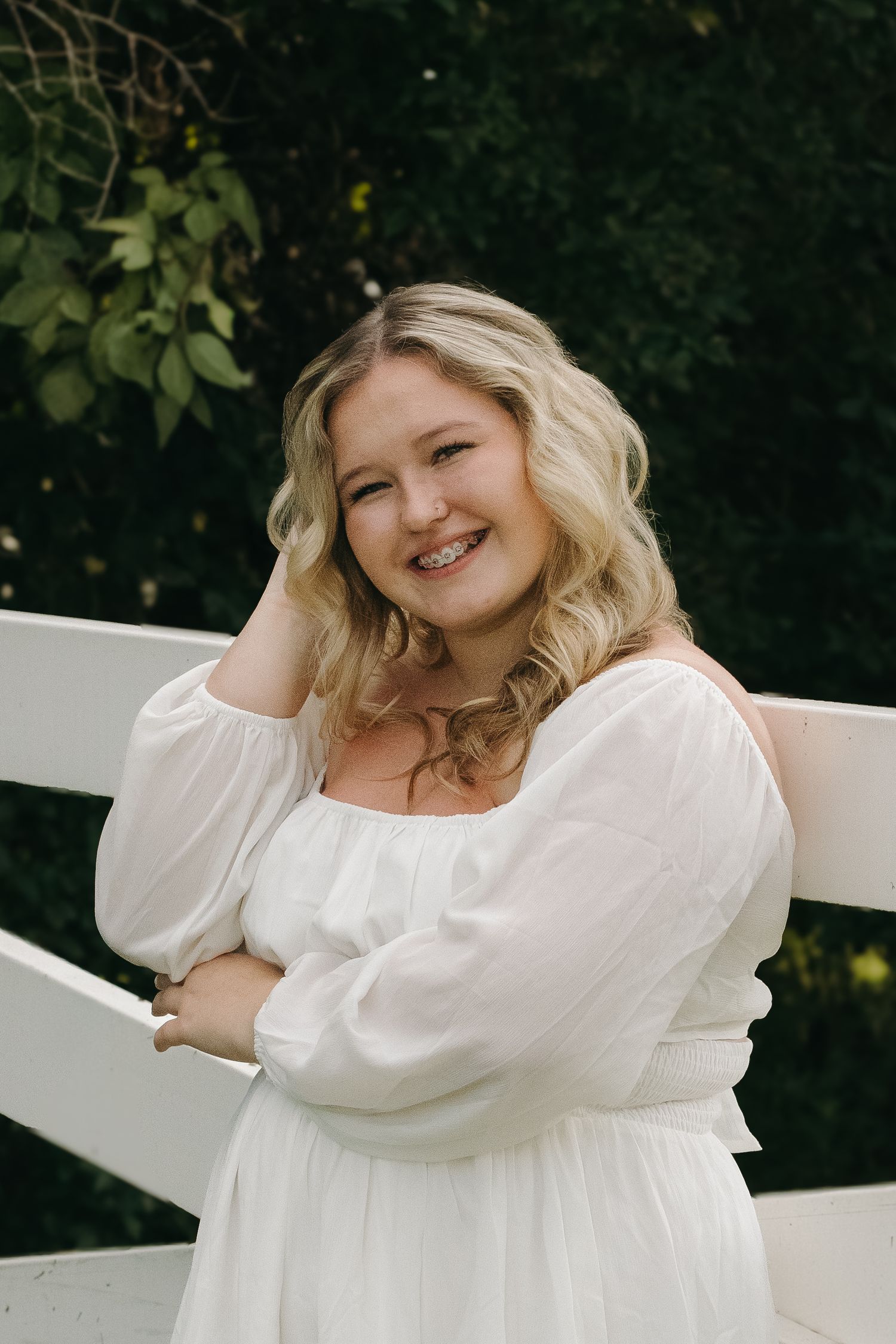Blonde woman in white dress smiles leaning against white fence with green foliage behind her.