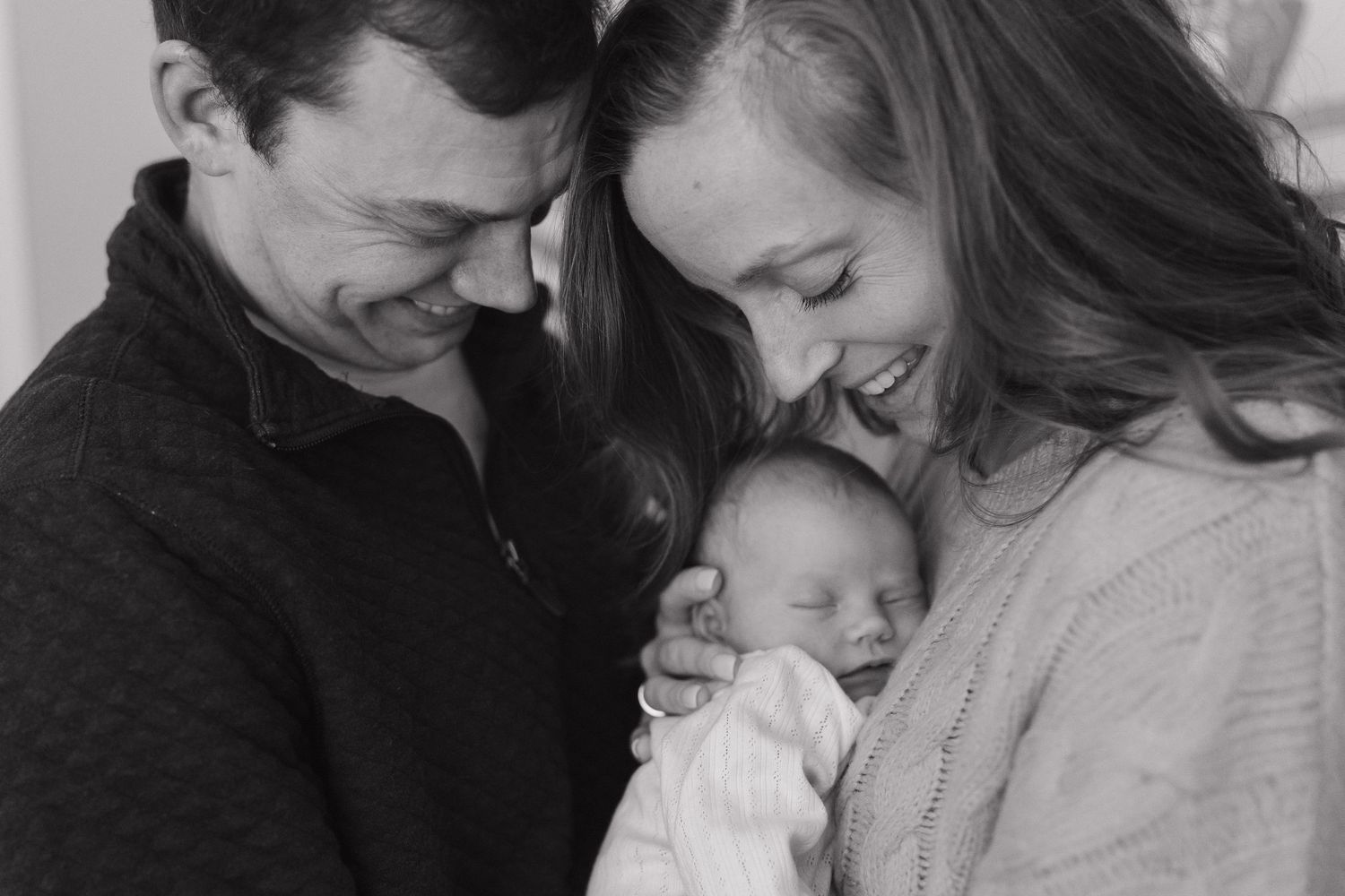 Black and white photo of smiling parents lovingly holding their newborn baby close together.