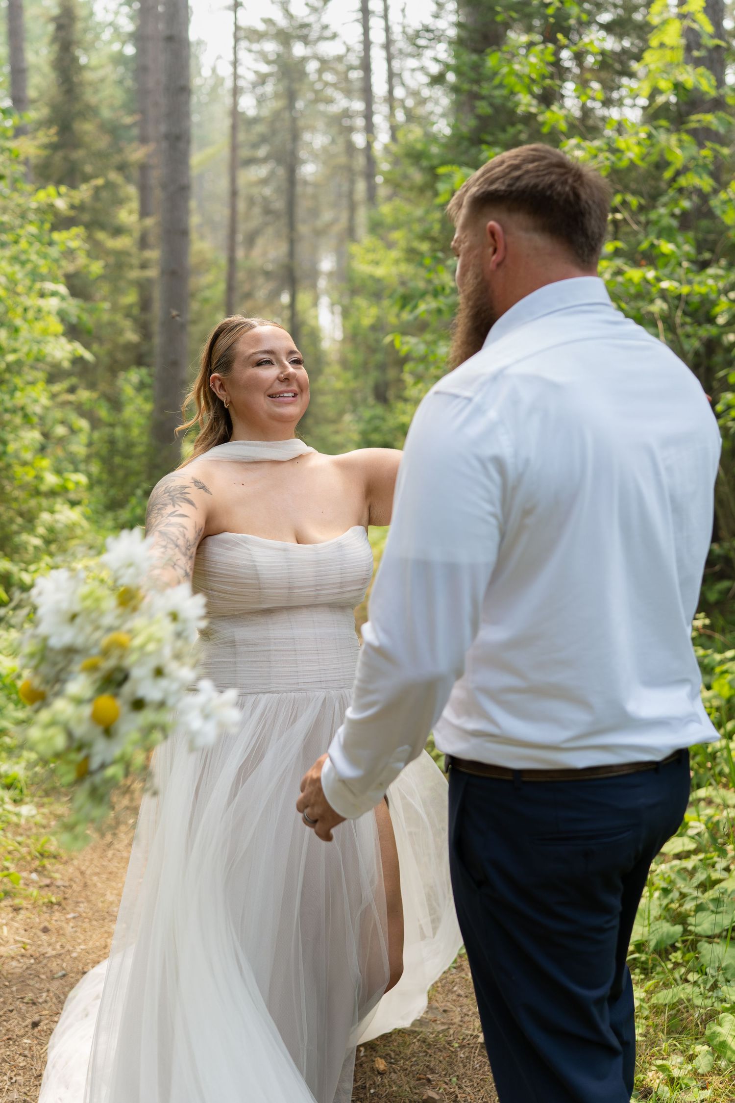 Bride in white gown holds groom's hand on forest trail, smiling joyfully during outdoor wedding ceremony.