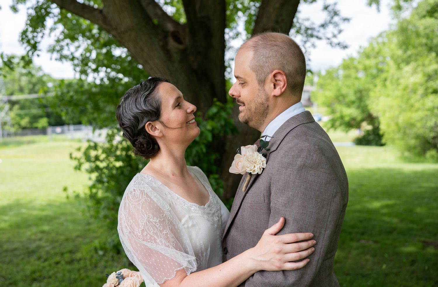 Bride and groom share a tender moment outdoors under a large tree on their wedding day.