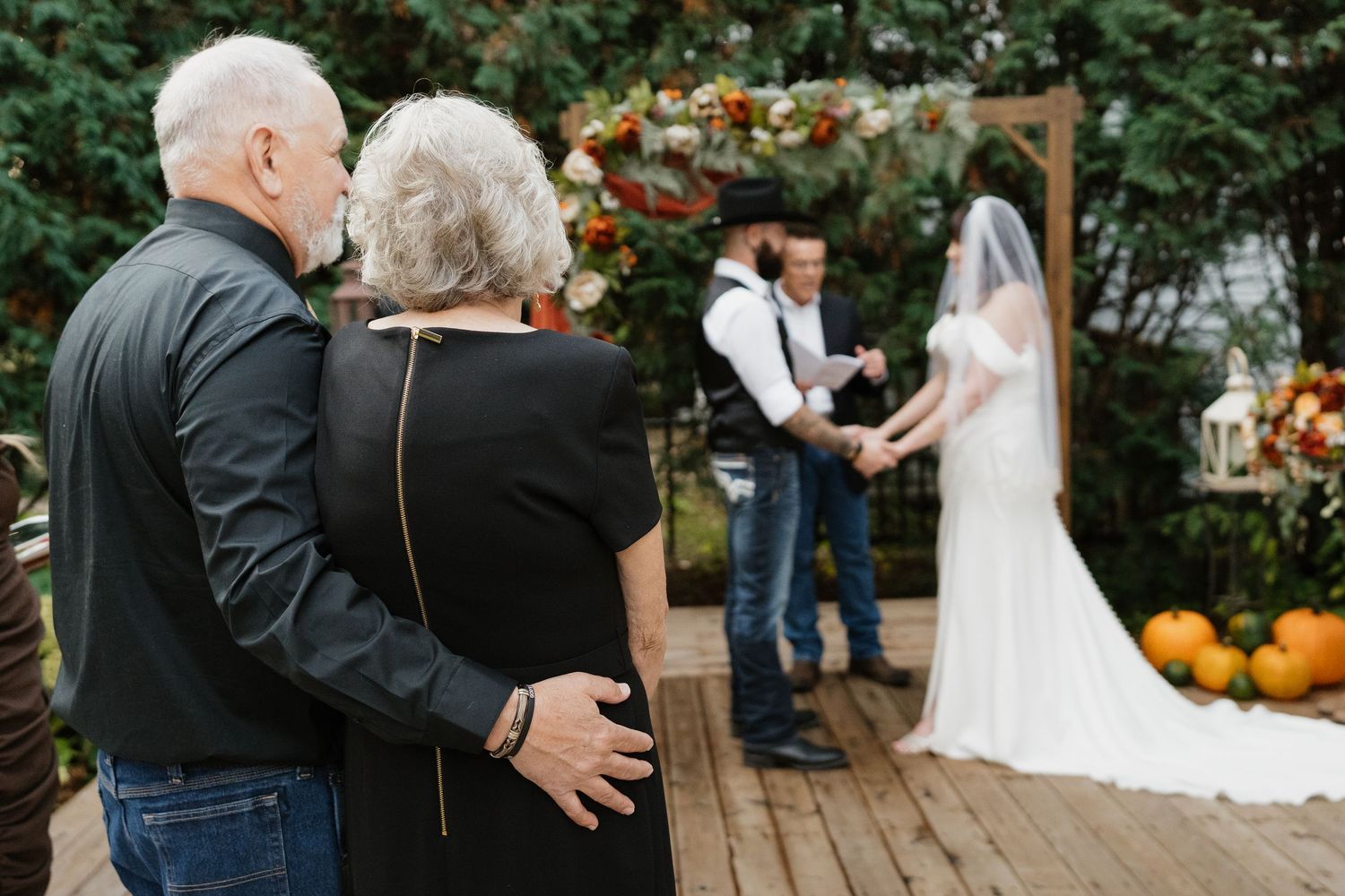 Elderly couple watches from behind as bride and groom exchange vows at outdoor ceremony.
