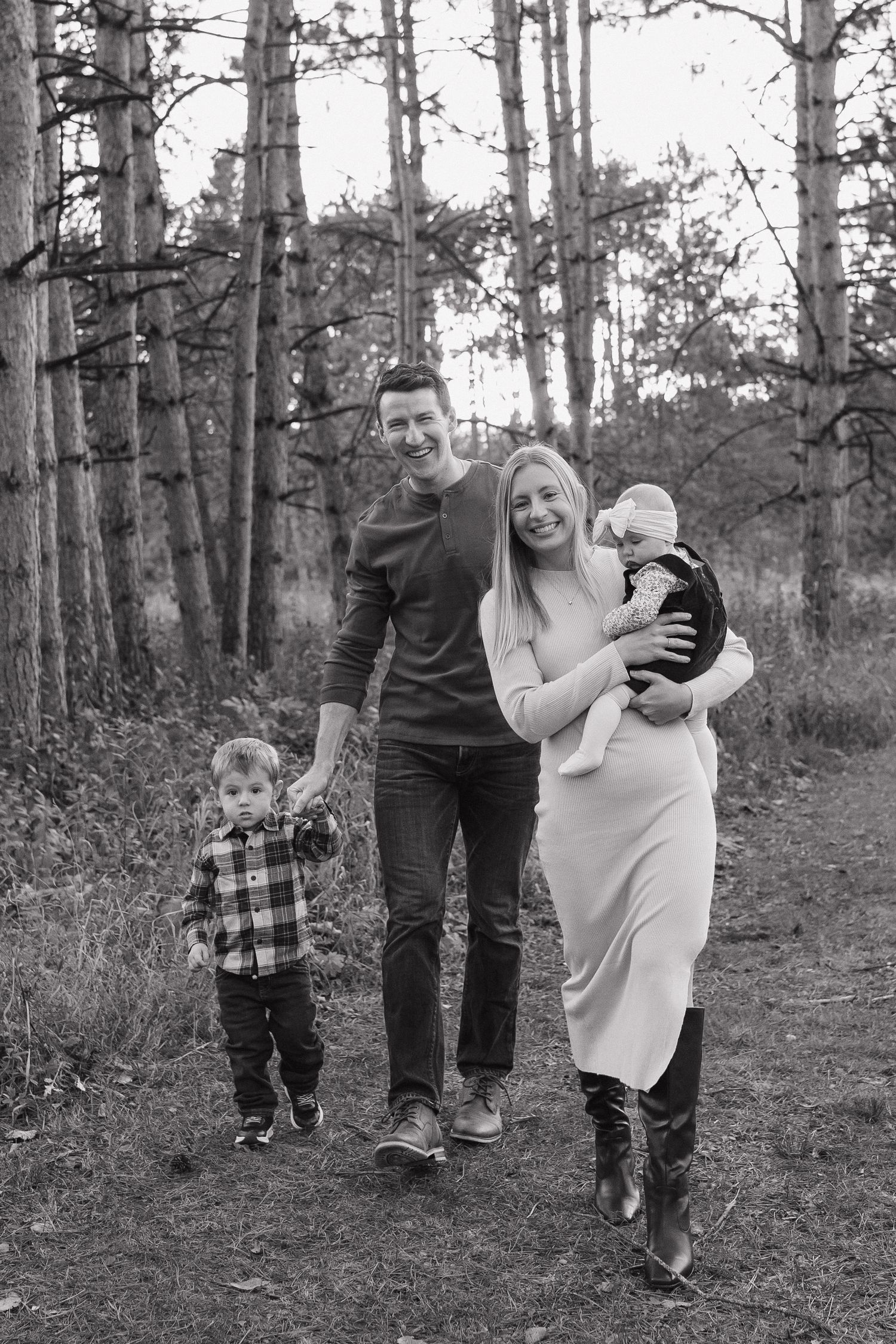 Black and white family portrait in forest, parents walking with toddler and baby in natural woodland setting.
