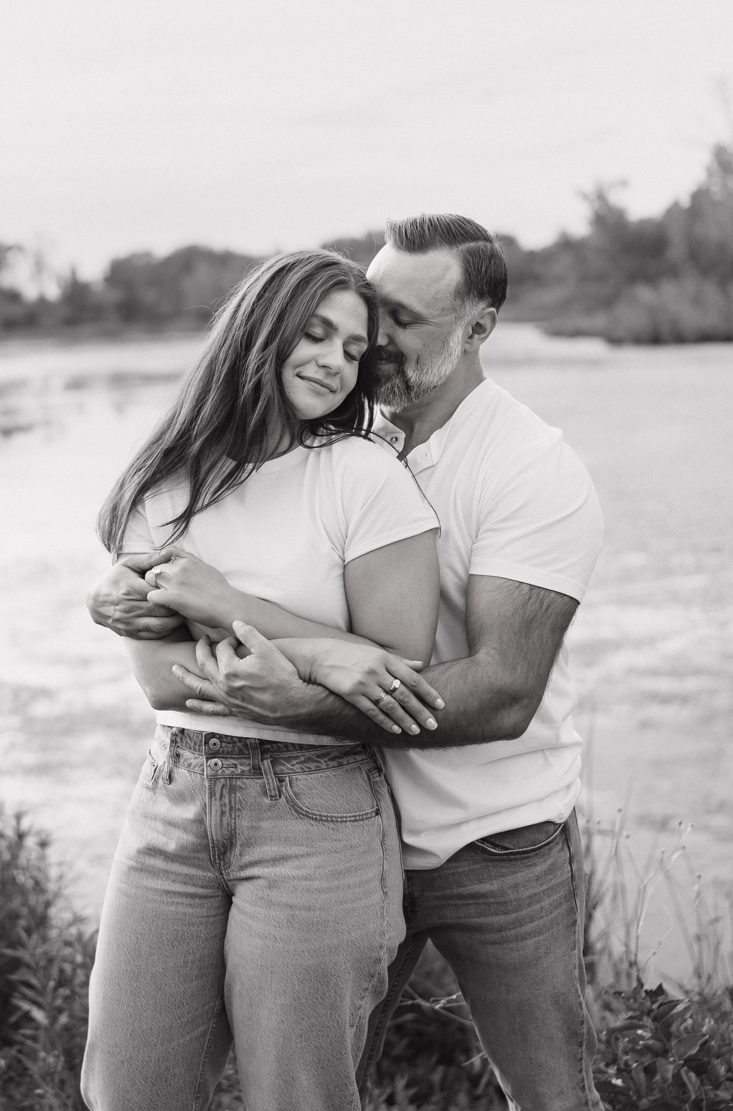 Black and white photo of a couple embracing near a river, man kissing woman's cheek while she smiles with eyes closed.