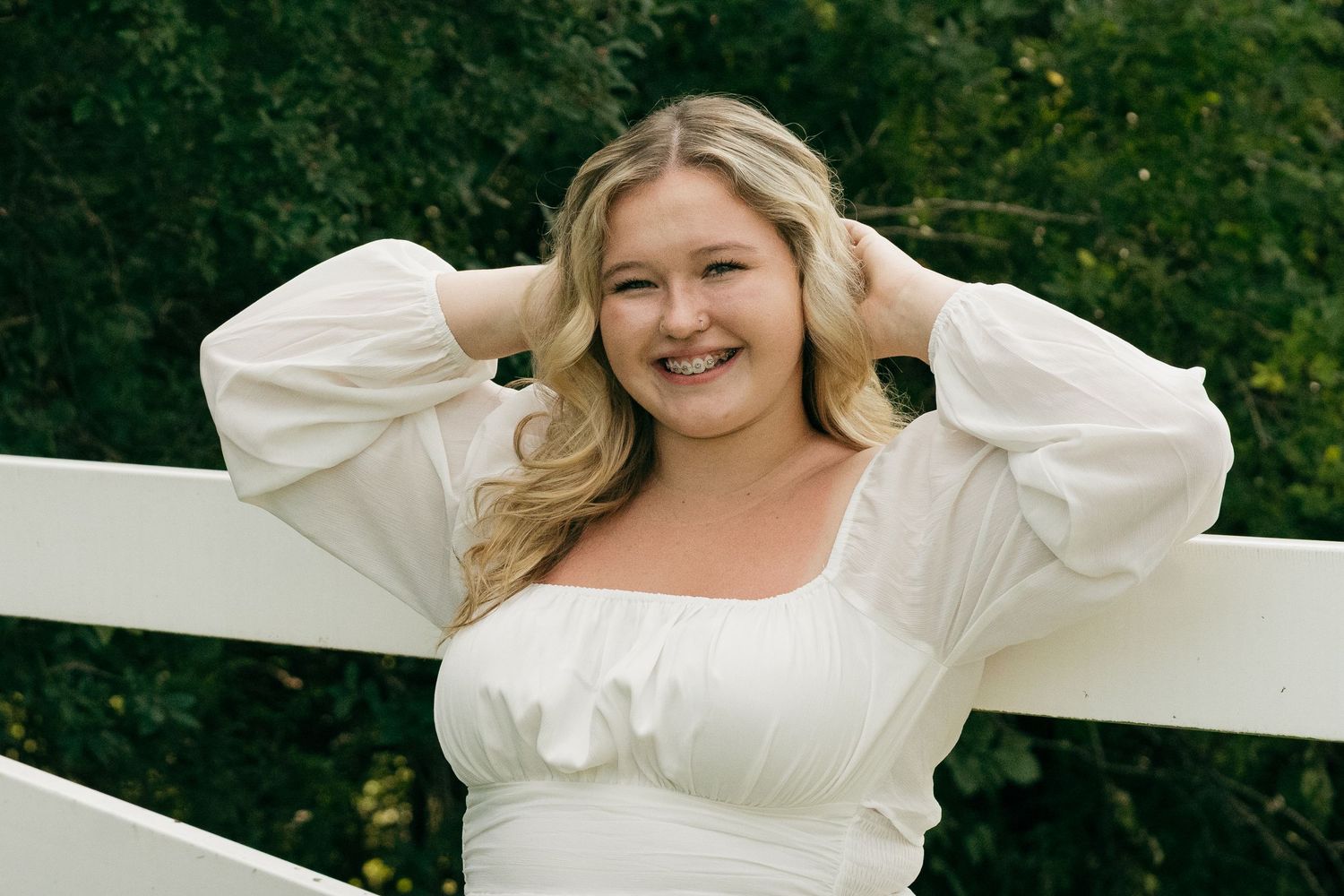 Cheerful blonde woman in white dress poses with hands in hair by white wooden fence.