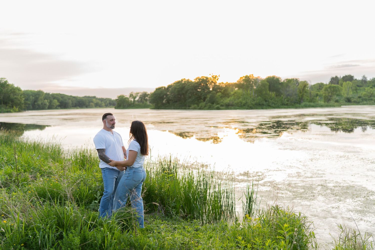 Couple holding hands by a serene river at sunset, surrounded by lush green grass and trees in a romantic outdoor setting.
