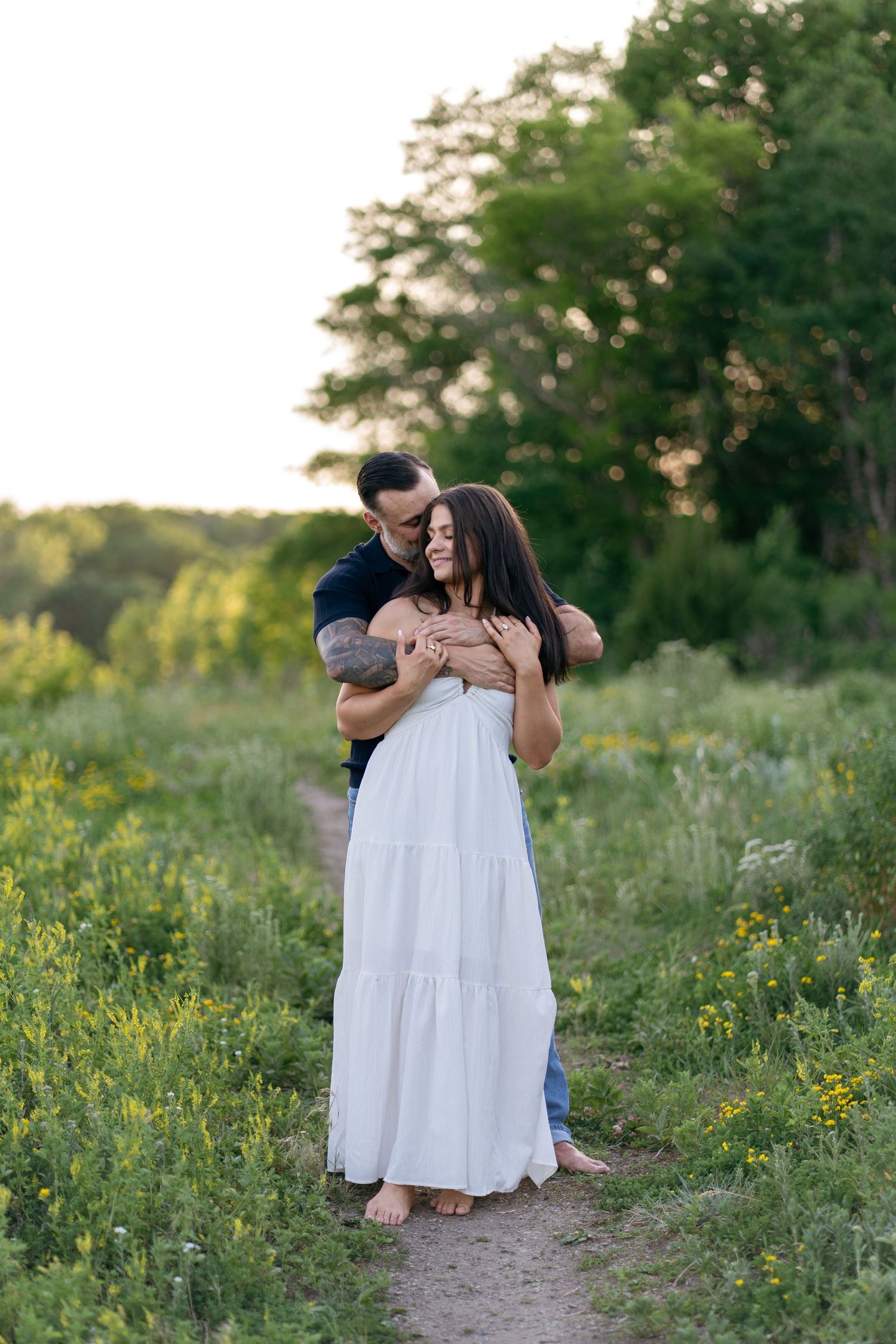 Couple embracing on a garden path, woman in white dress, man in dark shirt, surrounded by lush greenery at sunset.