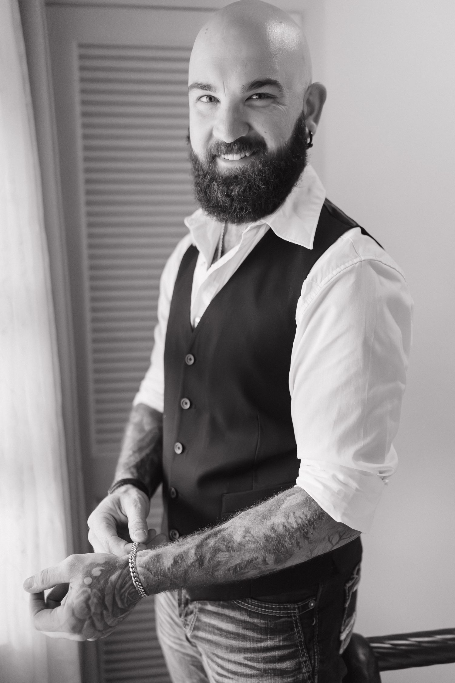 Black and white portrait of a bald bearded tattooed groom in a vest and white shirt.