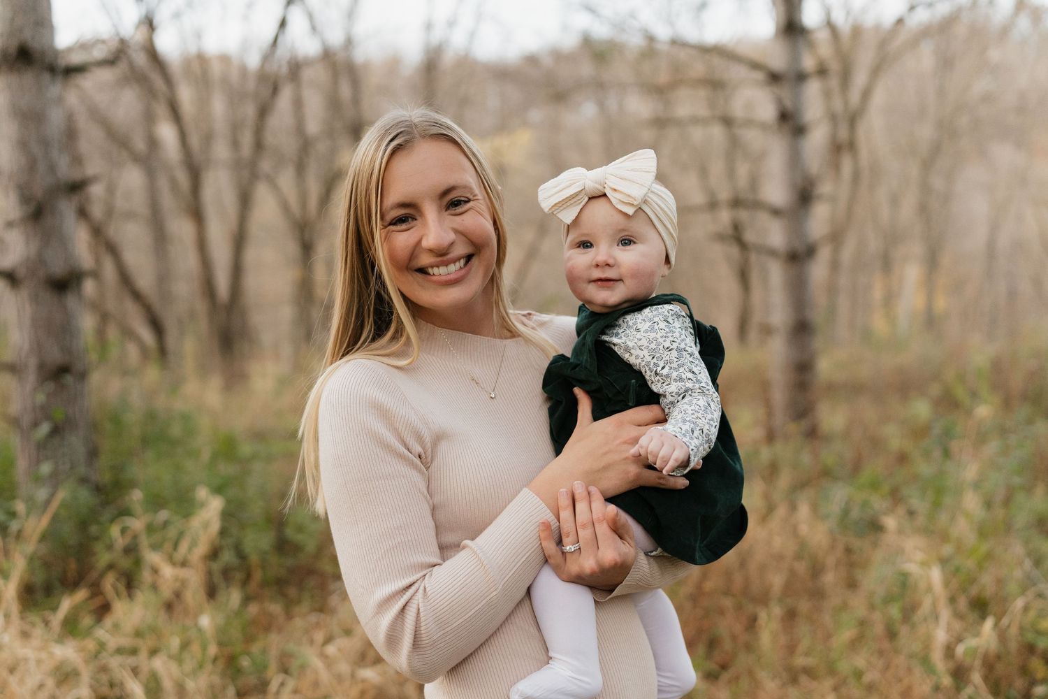 Smiling blonde woman in cream sweater holds baby girl in green dress and white bow headband outdoors.