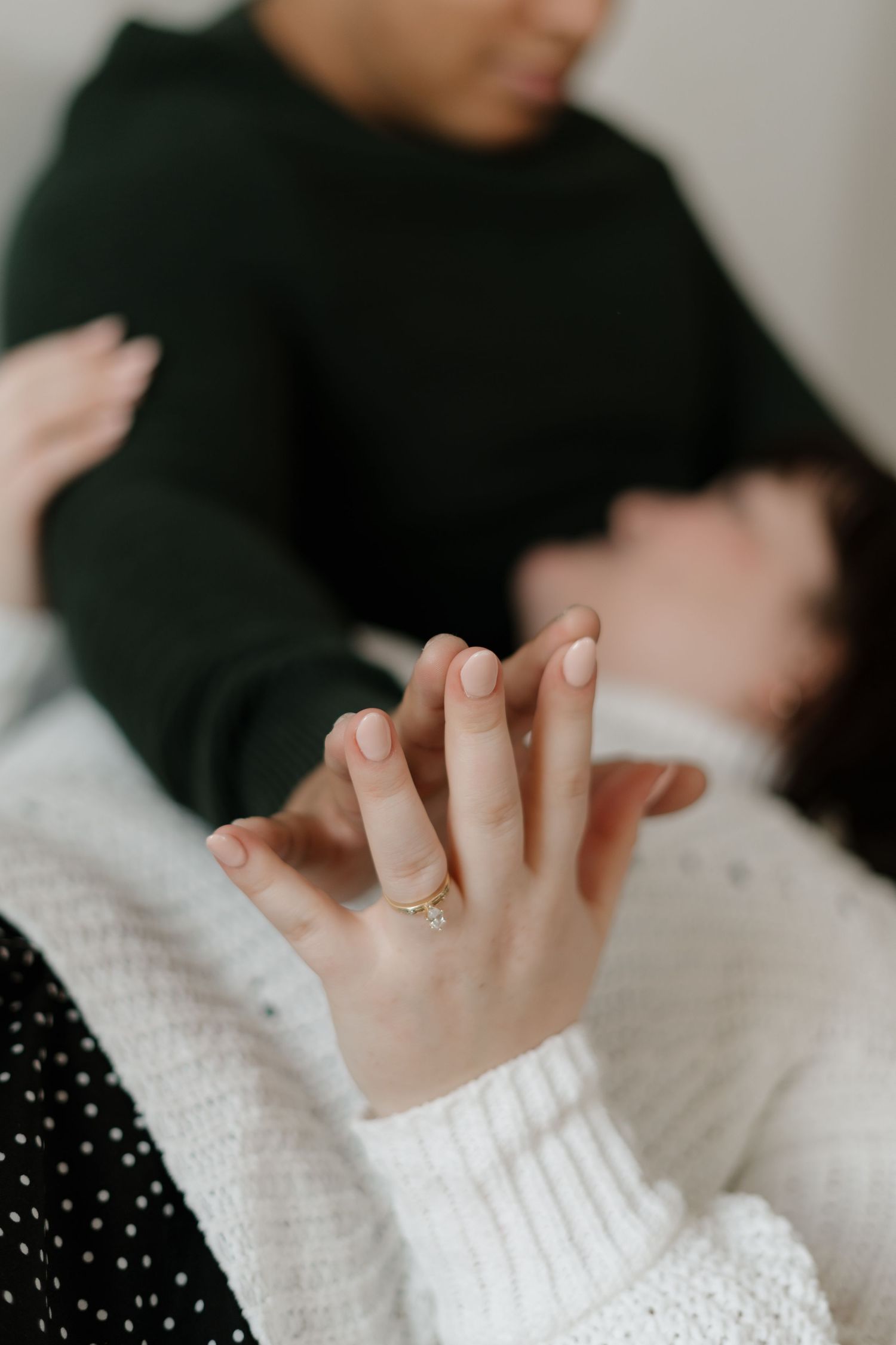Close-up of couple holding hands, woman wearing gold engagement ring, in soft focus romantic setting.