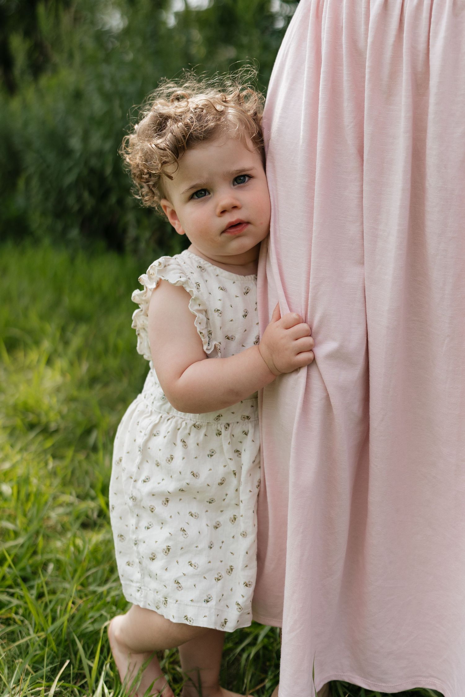 A curly-haired toddler in a white floral dress clings to her mothers pink dress outdoors.