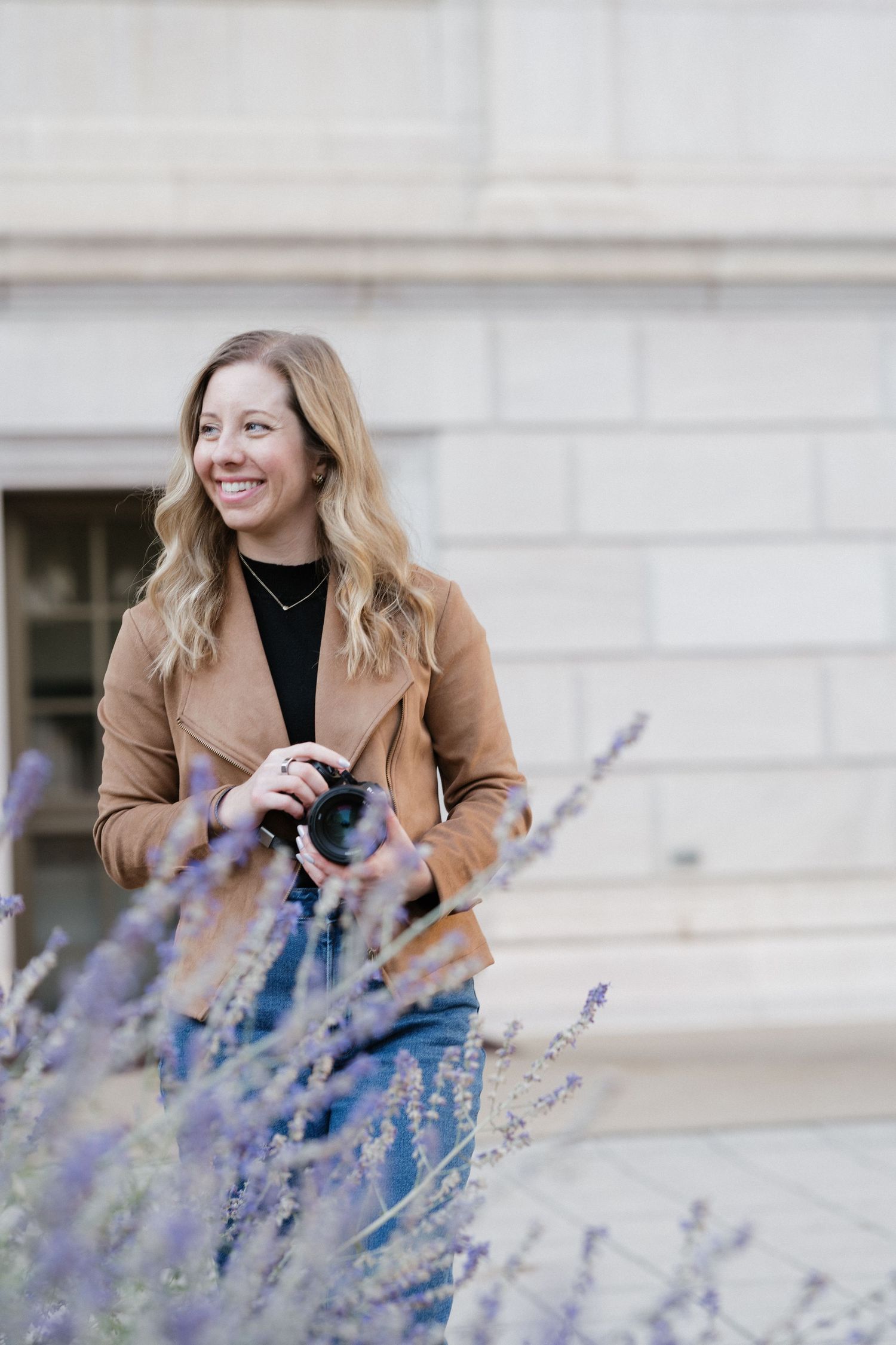 Cheerful female photographer with wavy blonde hair holding camera outdoors near purple lavender flowers.