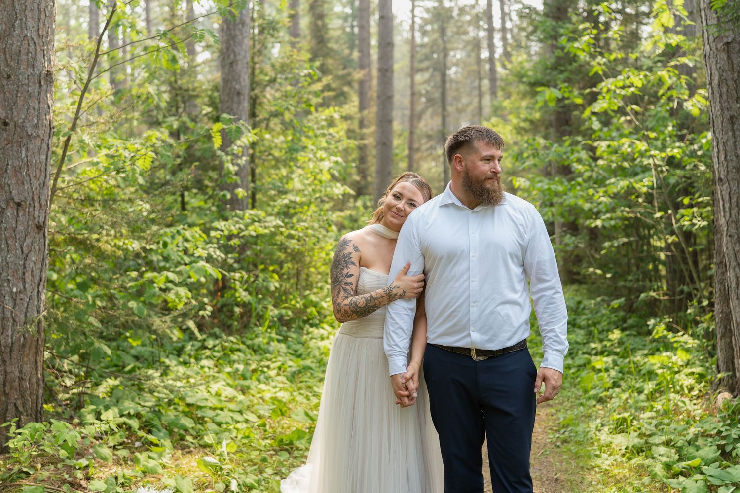 Bride and groom hold hands walking through a lush green forest in wedding attire, bride wearing a flowy white gown.