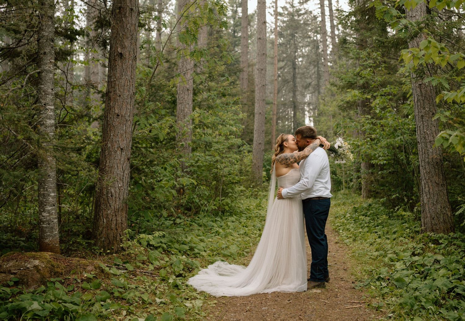 A bride and groom share a romantic kiss on a forest path surrounded by tall trees in a lush woodland wedding setting.
