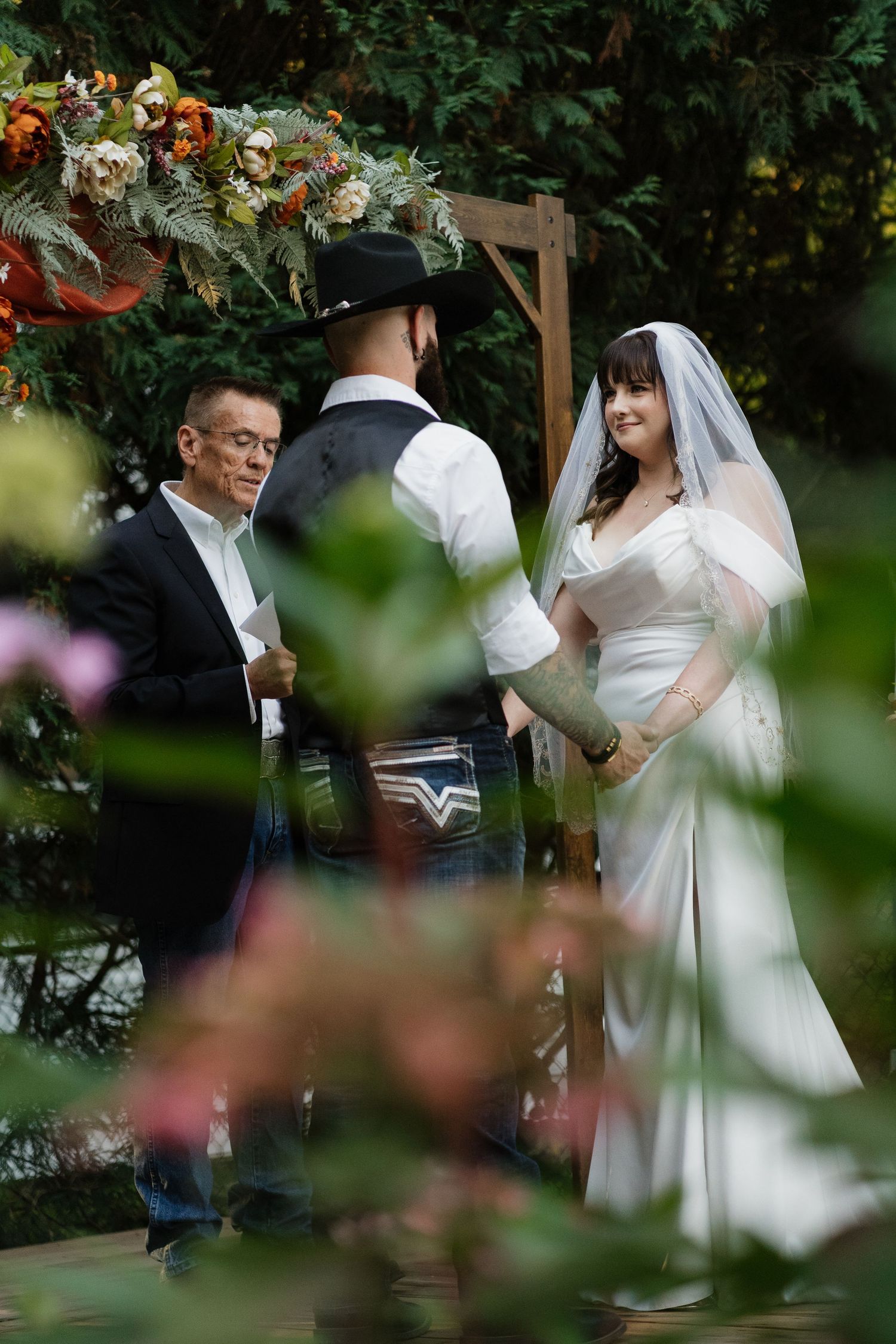 Bride in white gown and veil exchanges vows with groom in cowboy hat at outdoor garden wedding ceremony.