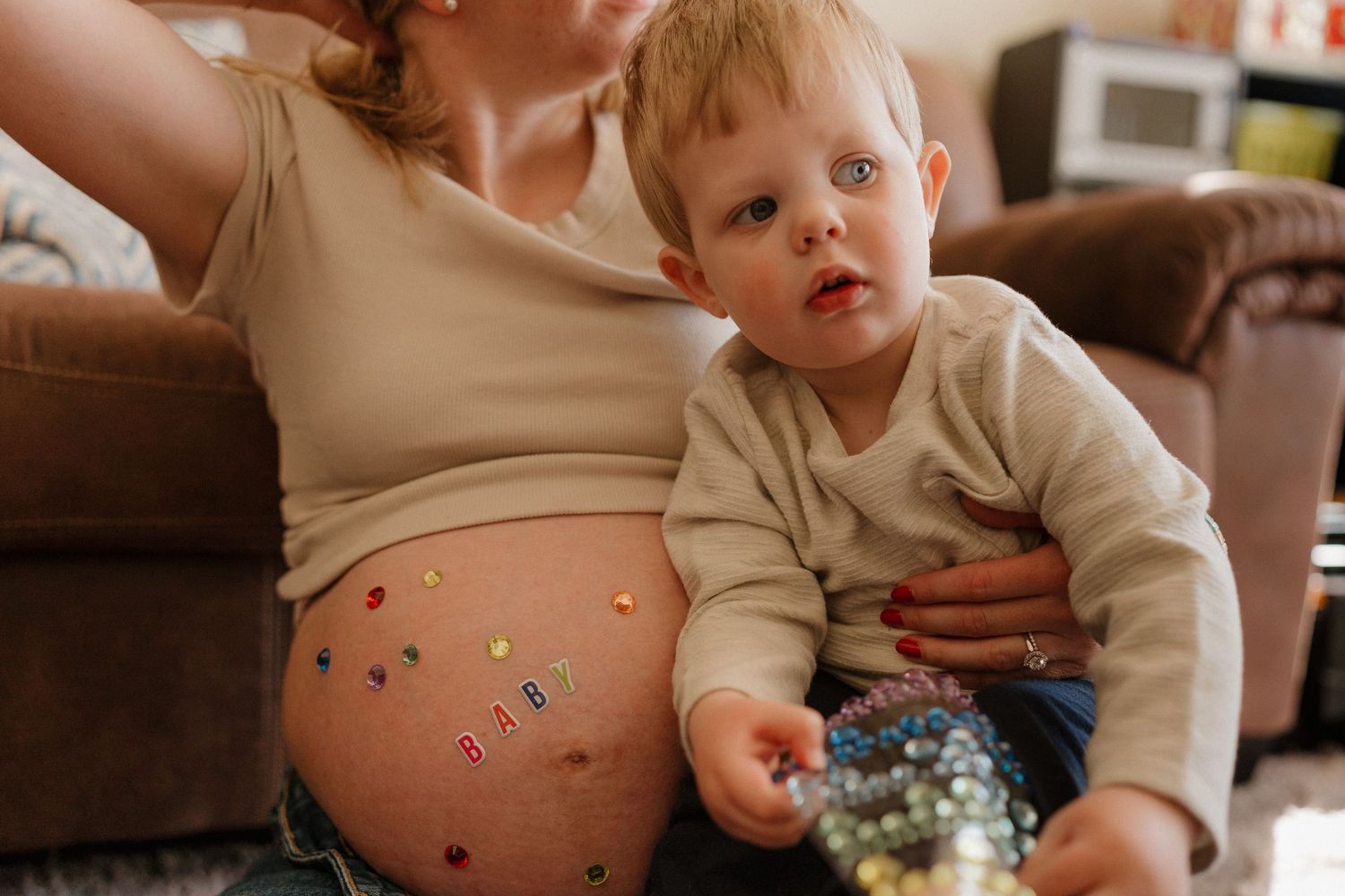 Pregnant woman with candy on belly sitting with toddler boy holding marbles on living room floor.