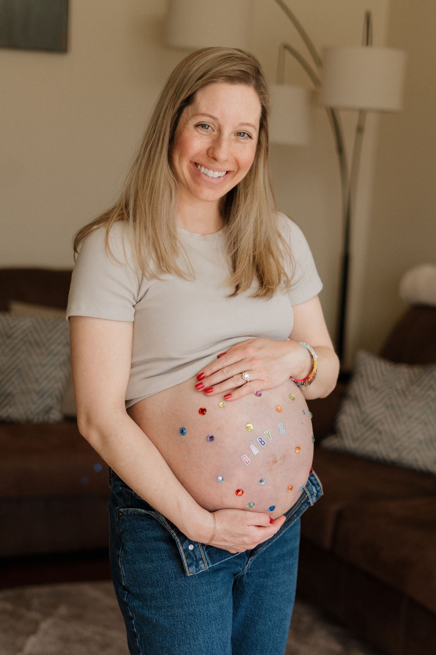 Smiling pregnant woman in gray crop top and jeans showing decorated baby bump with colorful stickers indoors.