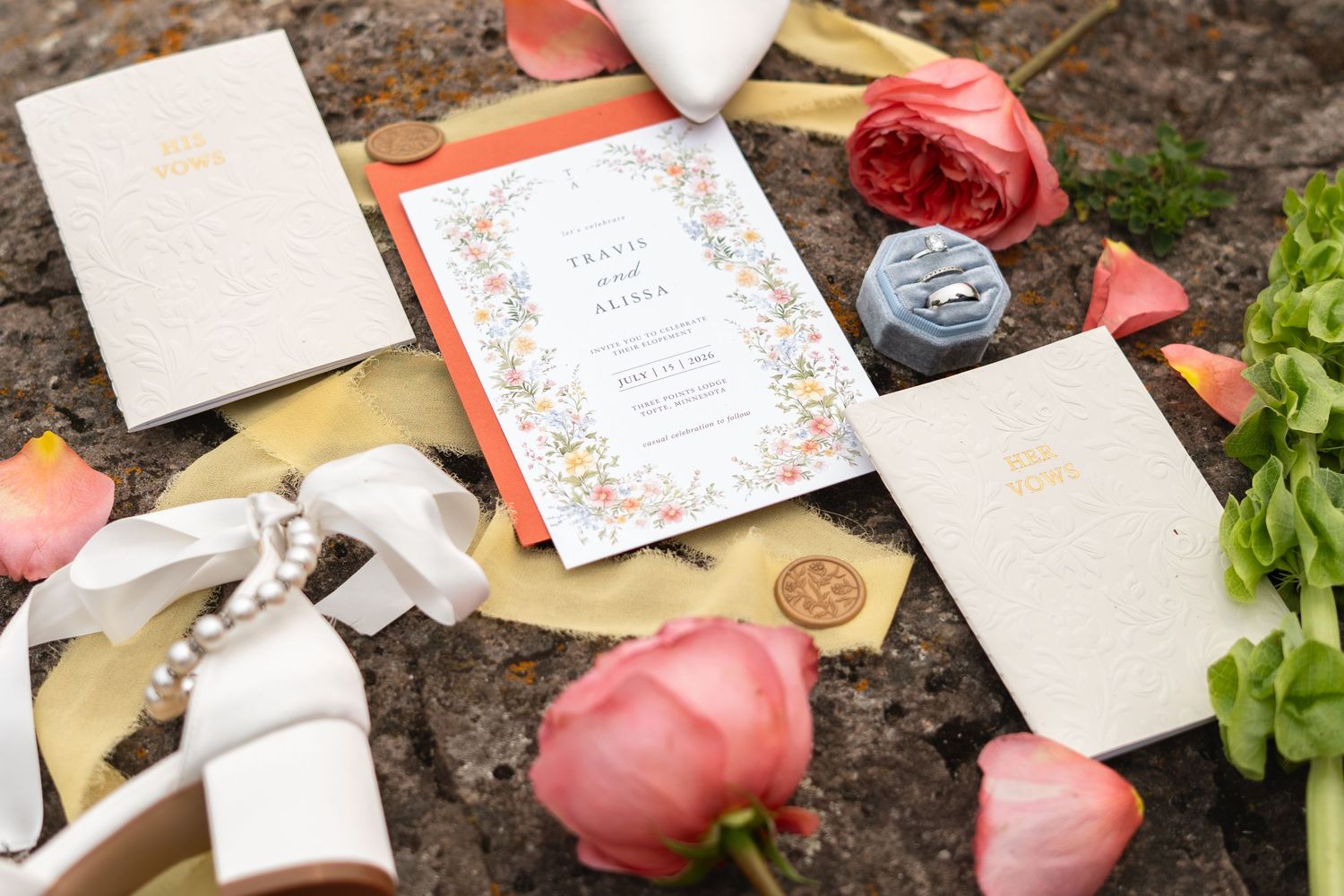 Elegant wedding invitation suite with floral border, pink roses, ribbon, and ring box on stone surface.