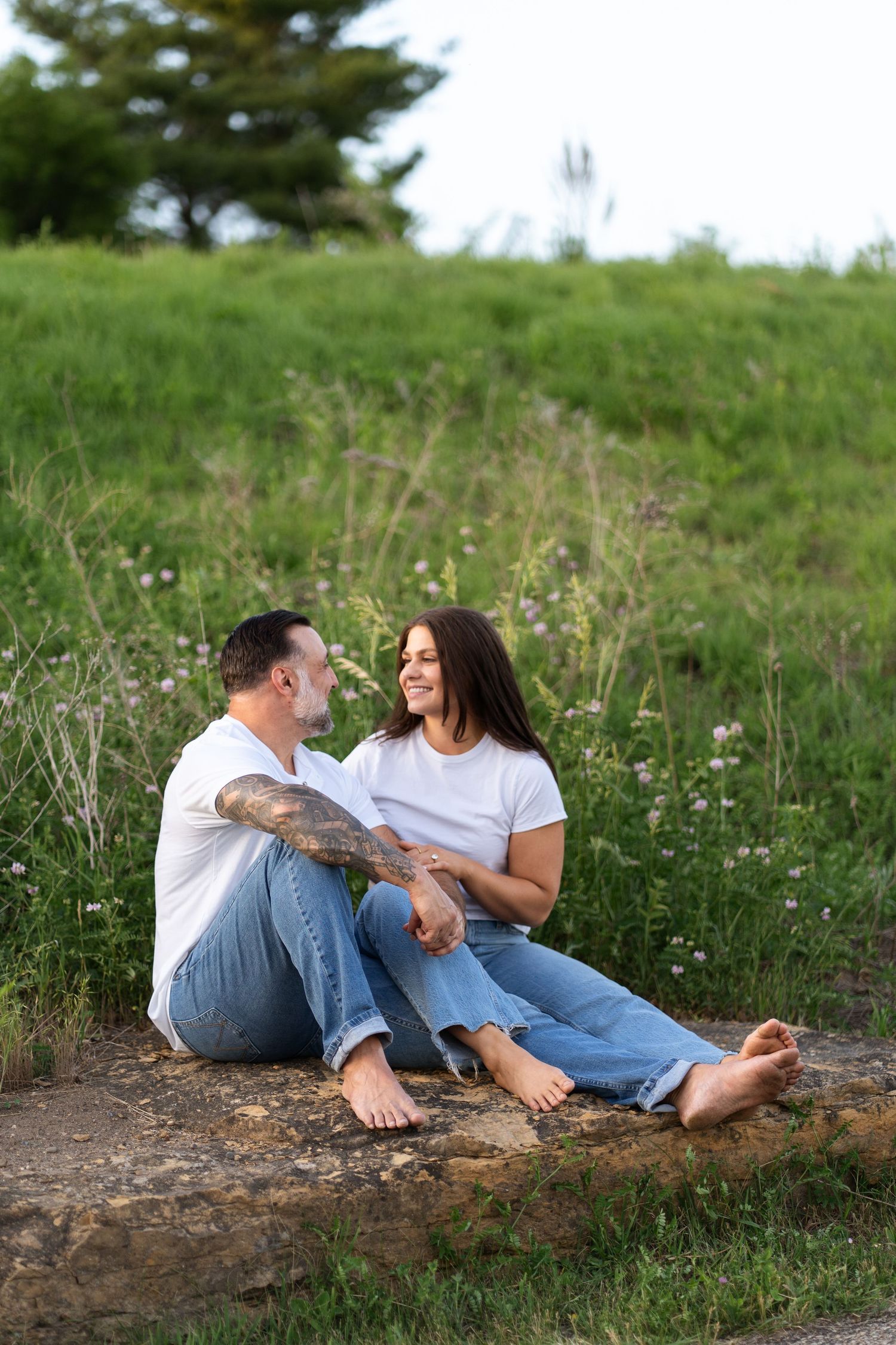 Couple in matching white tees and jeans sitting barefoot on a rock in a green meadow, smiling at each other.