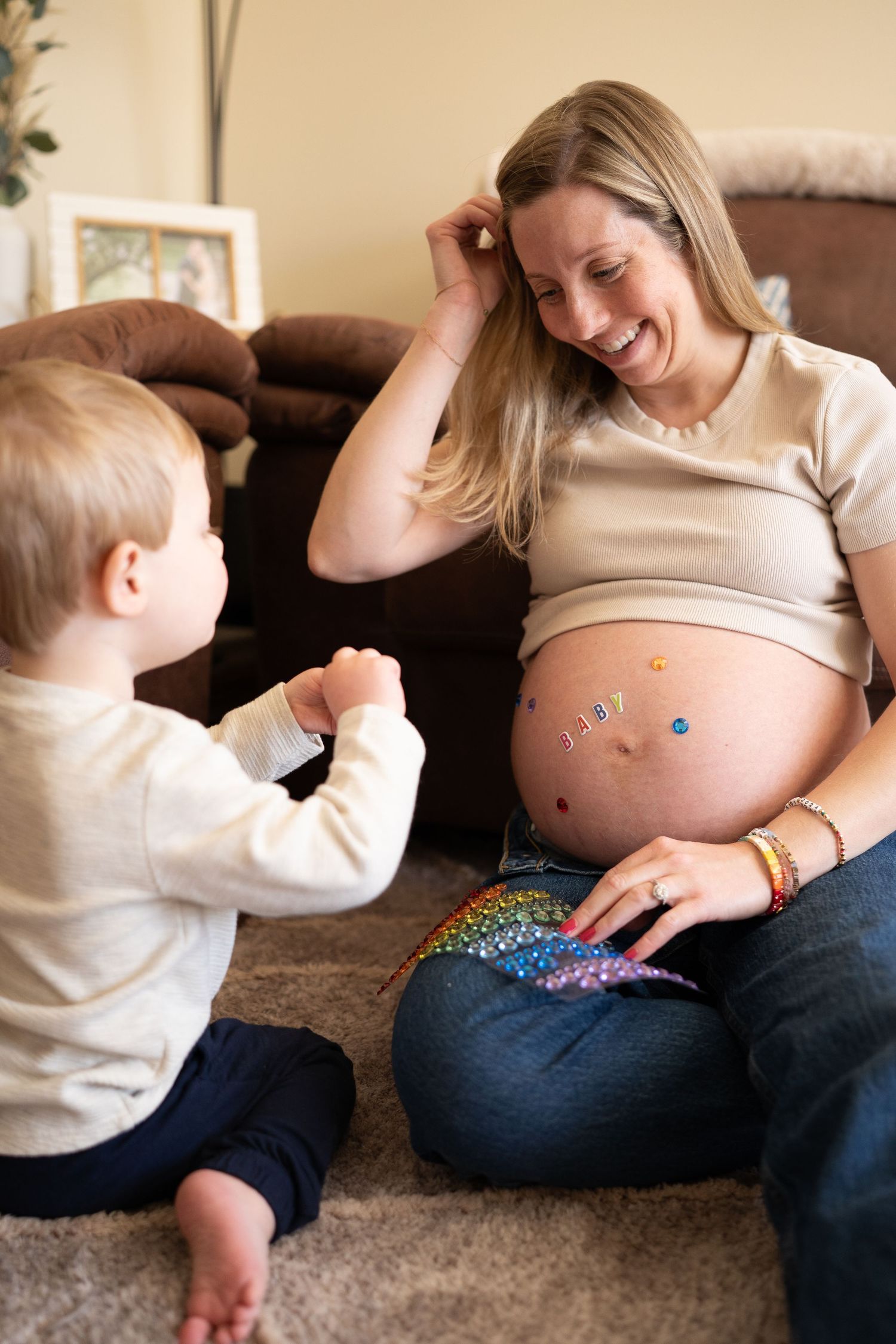 Smiling pregnant woman sits on floor with toddler, her decorated baby bump featuring colorful stickers and rainbow paint.